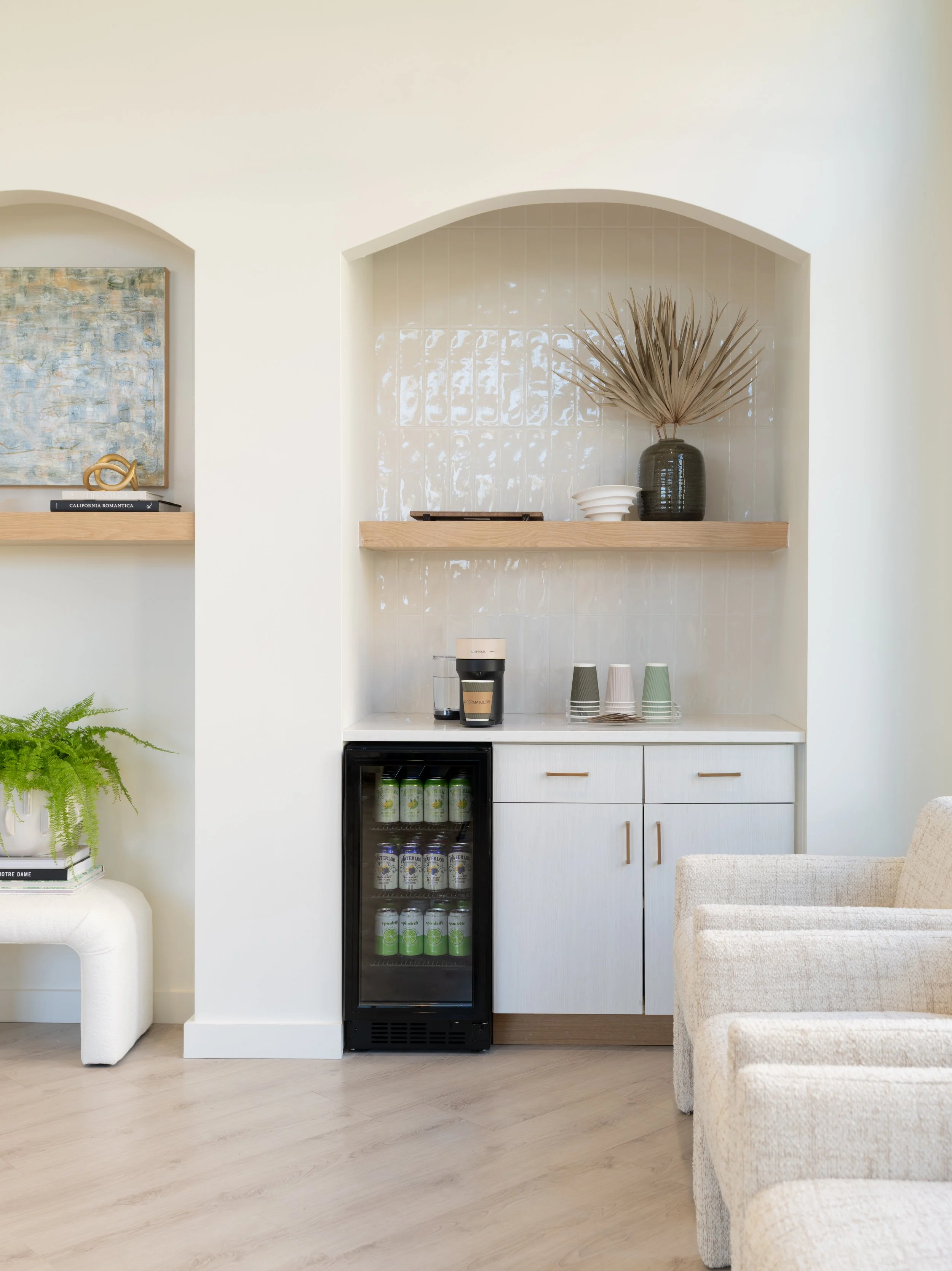 Interior kitchen nook with white cabinets, mini fridge with drinks, coffee machine, and decorative vase with dried palm leaves, beige armchairs, and a green fern on a white side table.