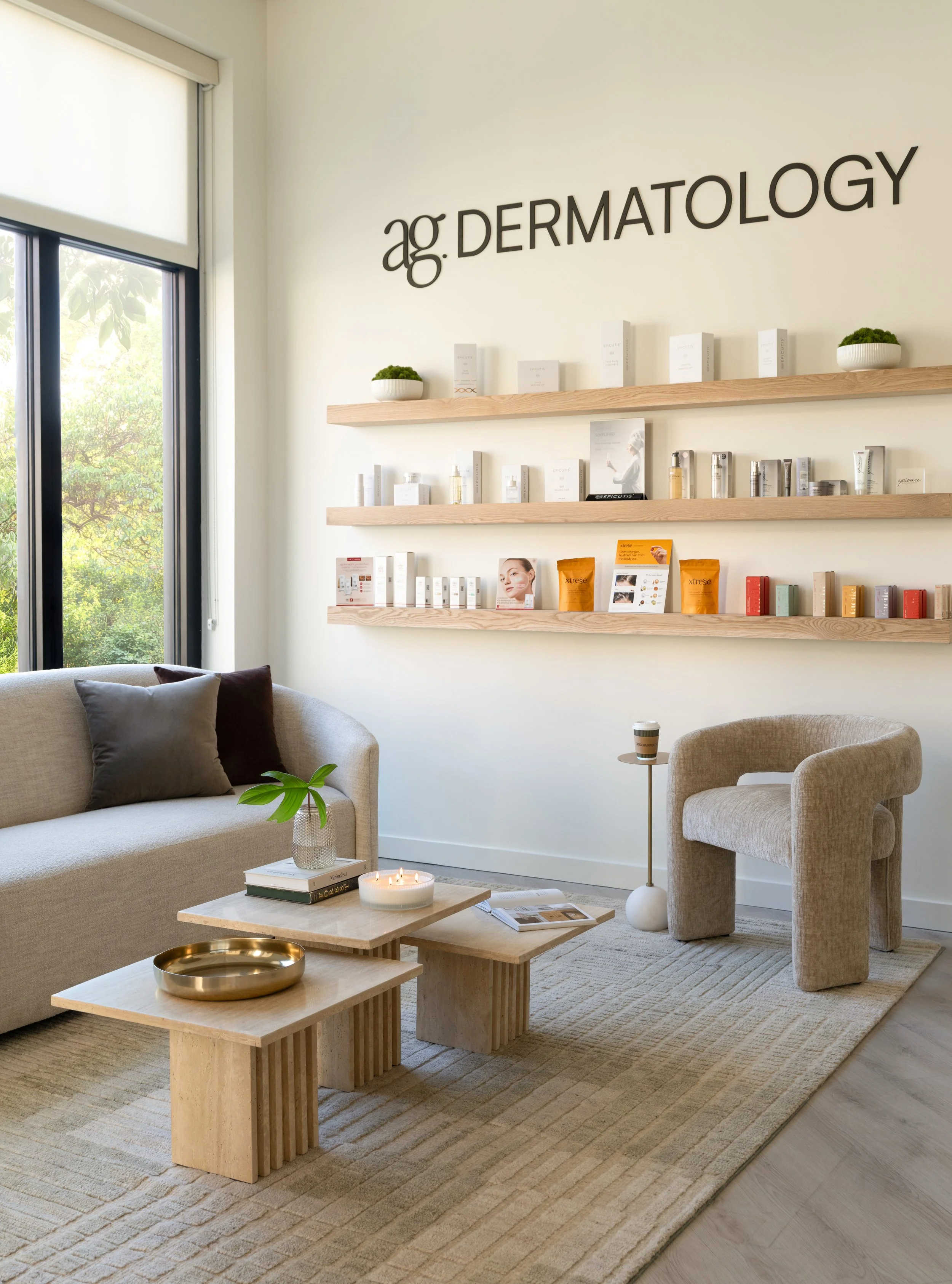 A dermatology clinic waiting area with beige couch, armchair, wooden tables, potted plants, wall-mounted shelves with skincare products, and large window with greenery outside.