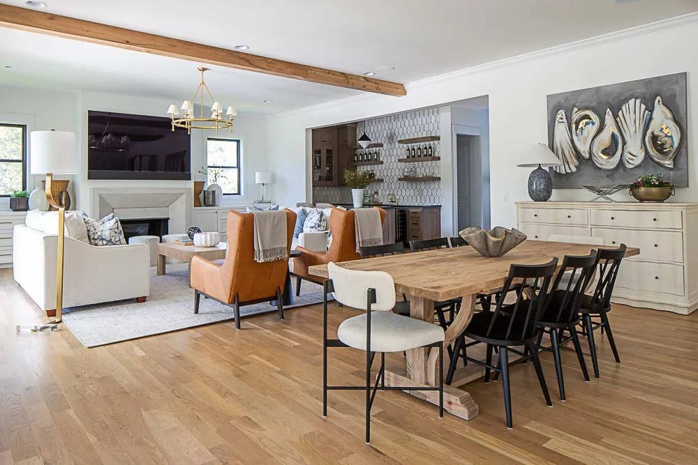 Modern open-concept living and dining area with a wooden dining table, black chairs, beige sofa, orange armchairs, and a fireplace. Decor includes a chandelier, abstract wall art, and hardwood floors.