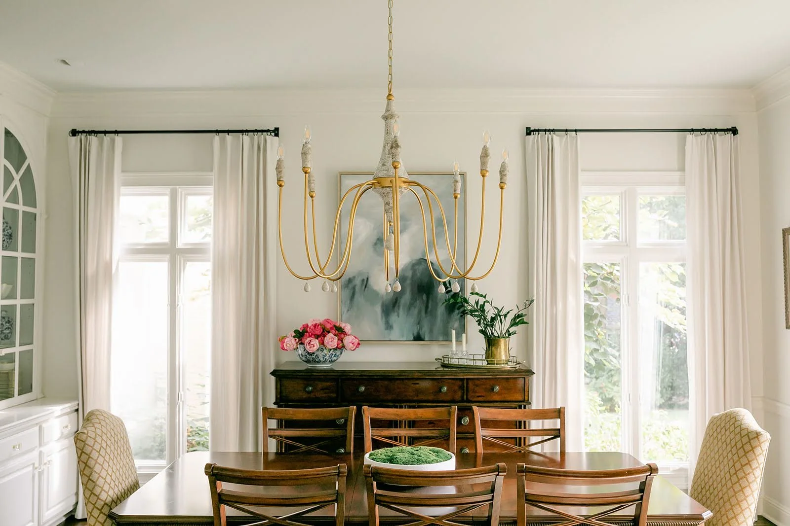 Elegant dining room with large wooden table, cushioned chairs, and a golden chandelier. A sideboard with pink flowers and a plant underneath a painting. Bright windows with white curtains line the back wall.