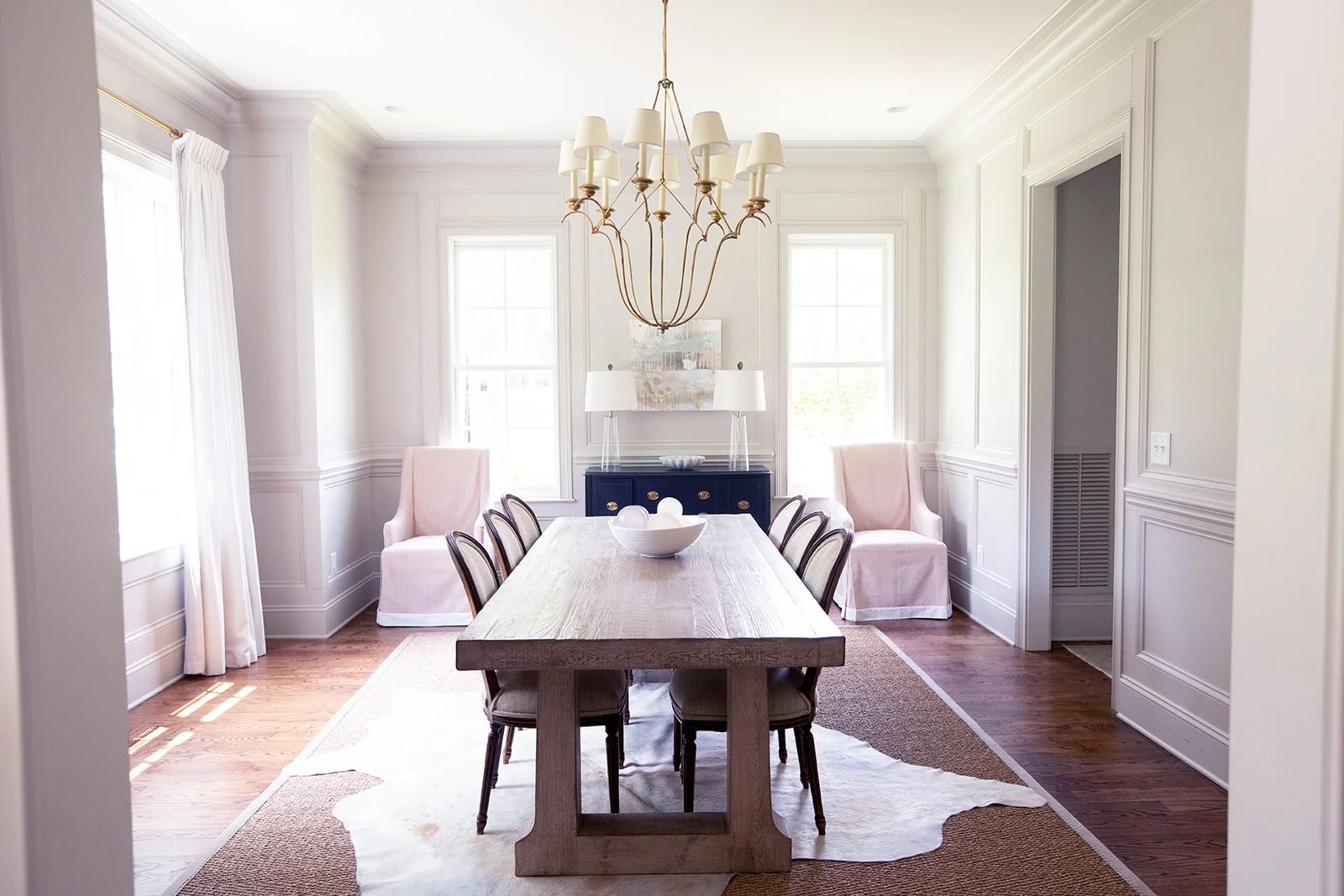 Elegant dining room with a large wooden table surrounded by chairs, a chandelier overhead, white walls, large windows, and minimalist decor featuring a blue sideboard with lamps.