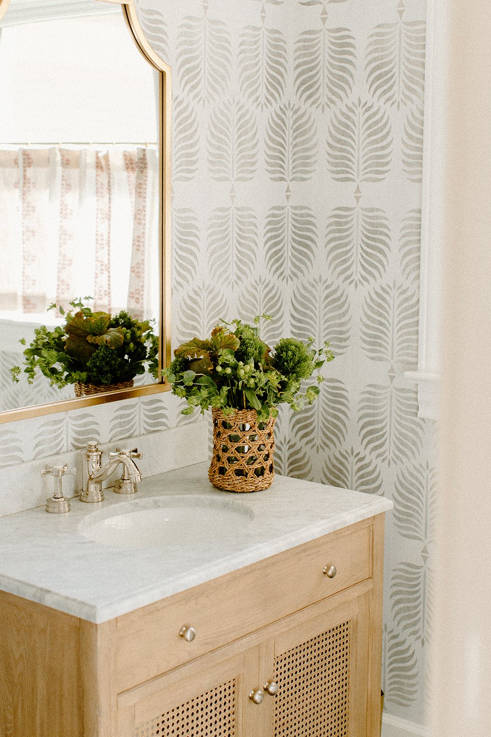Bathroom vanity with marble countertop, wicker plant holder, leafy greens, decorative mirror, and leafy wallpaper pattern.