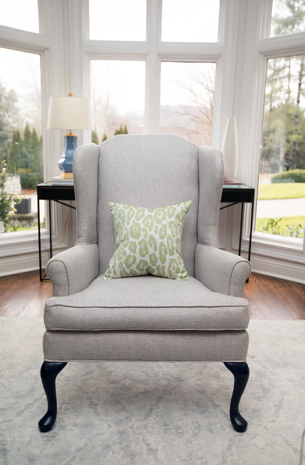 Gray upholstered wingback chair with a green patterned pillow, sitting in a room with large windows and a lamp on a side table.