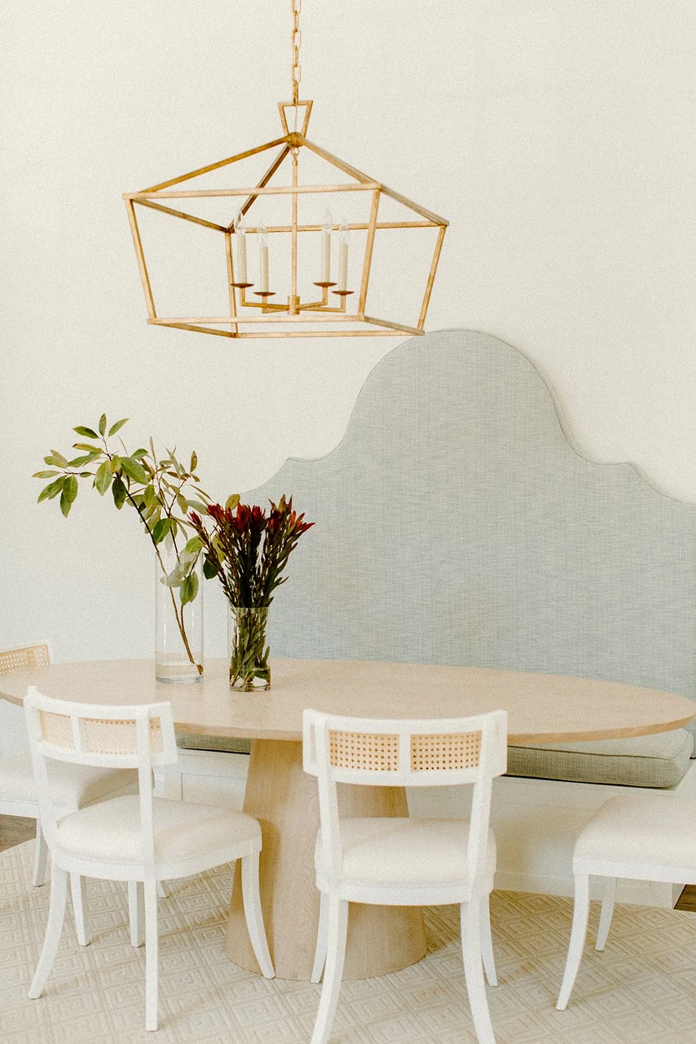 Dining area with a round wooden table, white chairs with cane backs, a geometric gold pendant light, and a floral arrangement in glass vases.