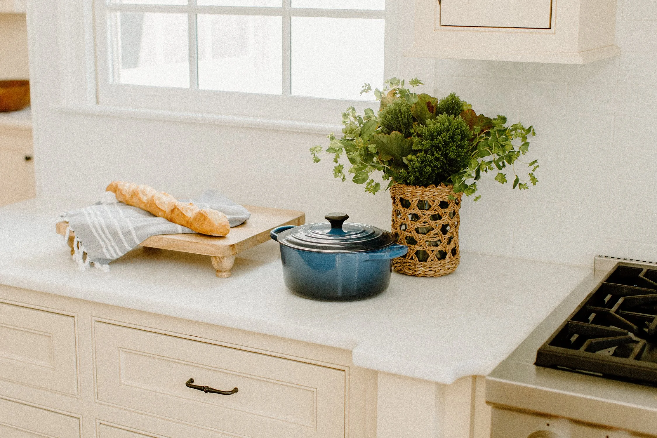 Cozy kitchen scene with a cutting board holding a baguette next to a blue Dutch oven and a basket of fresh herbs on a white countertop.