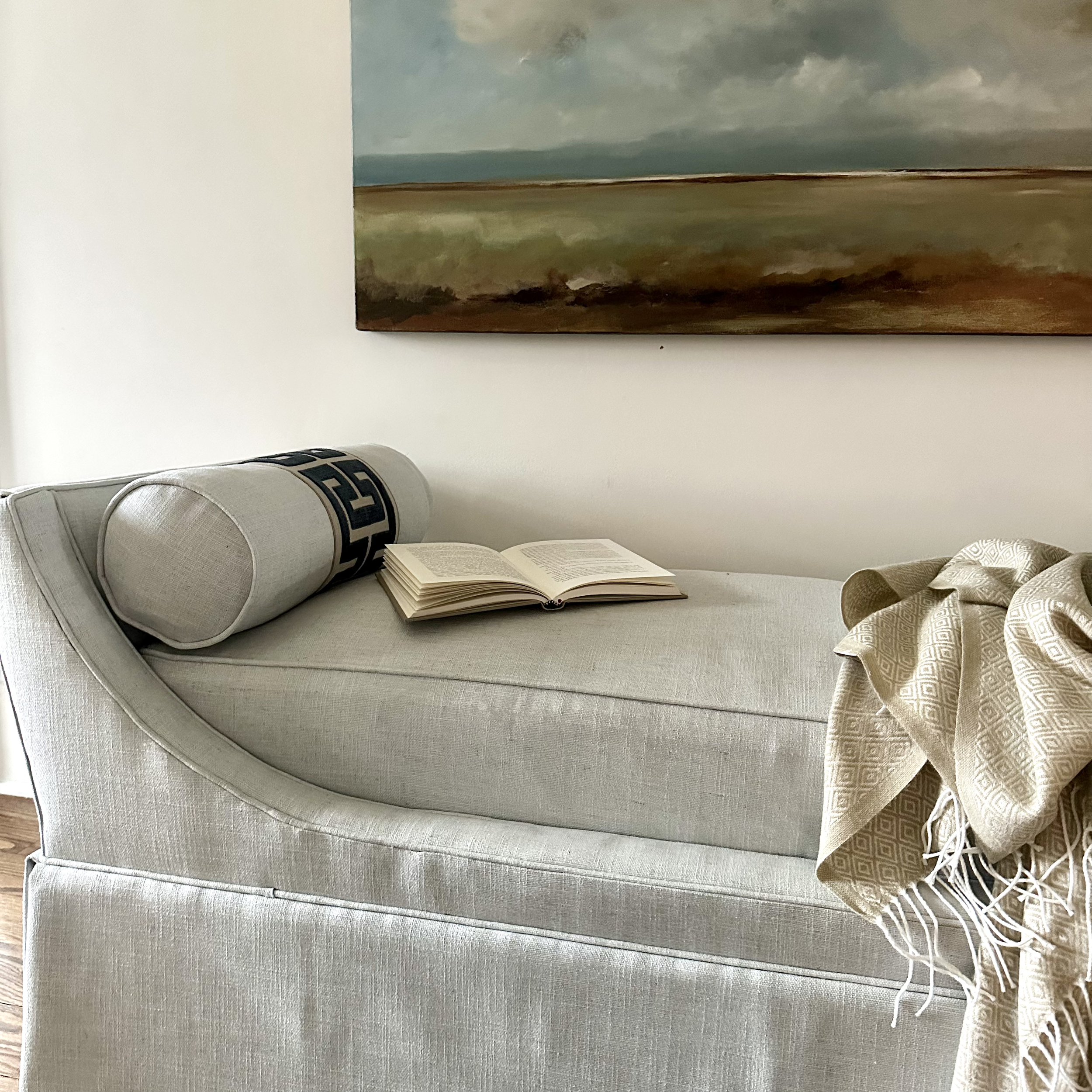 Elegant chaise lounge with a book and a throw blanket, featuring a landscape painting on the wall.