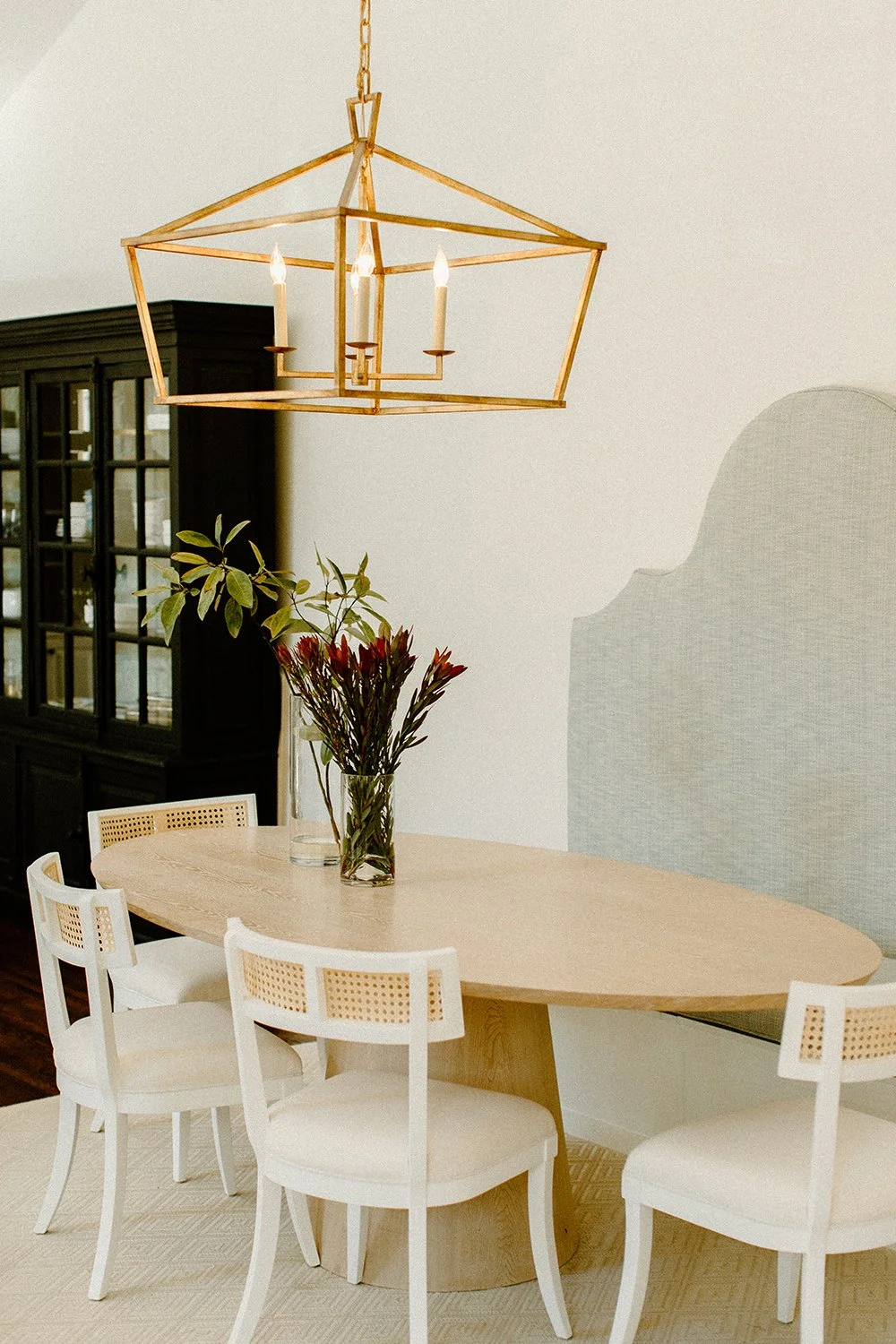 Dining room with oval wooden table, white chairs, a glass vase with flowers, a gold geometric chandelier, and a dark cabinet in the background.