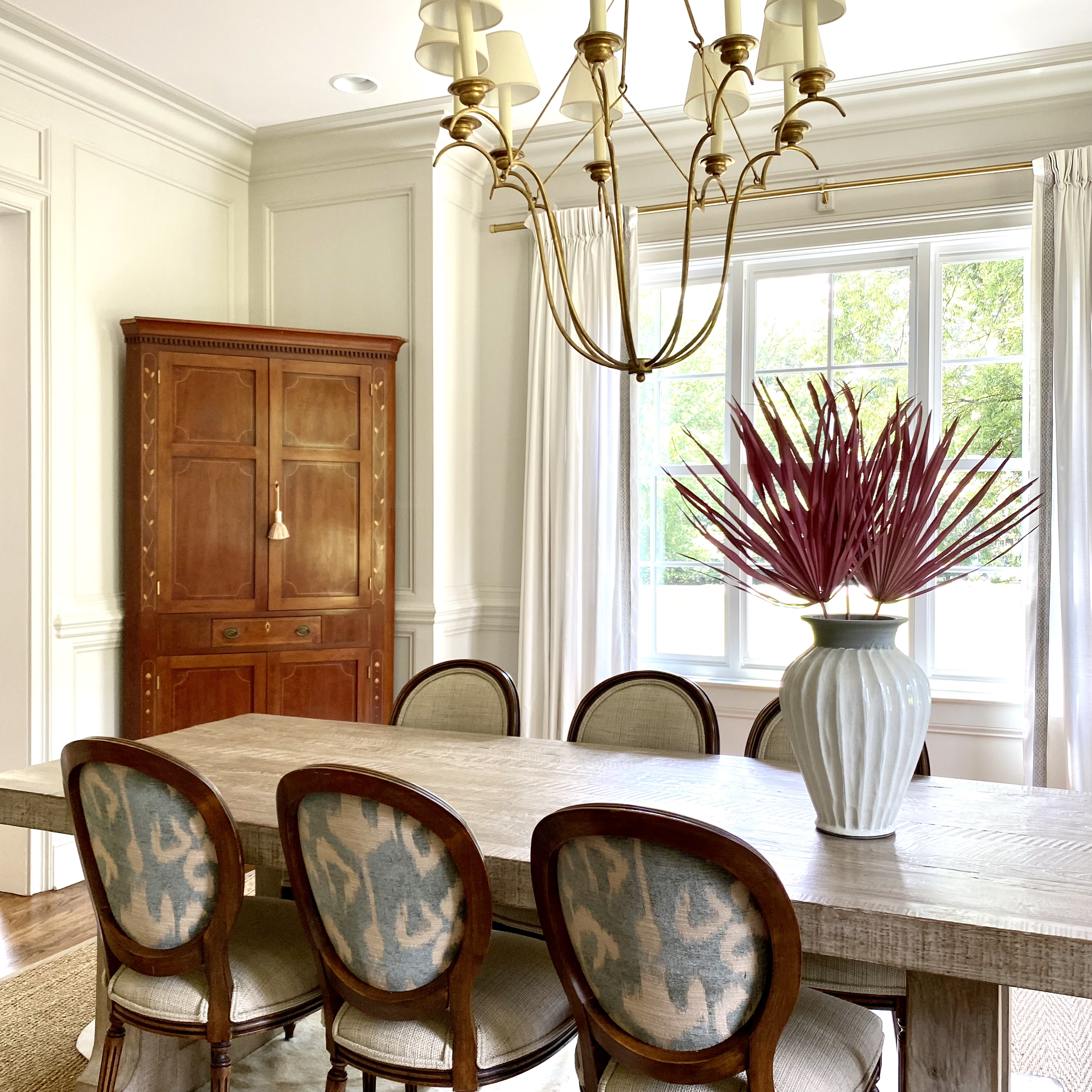 A dining room with a wooden table and six upholstered chairs featuring patterned backs. A large white vase with decorative red foliage is placed on the table. A chandelier hangs above. A wooden cabinet is against the wall, and a window with white cur