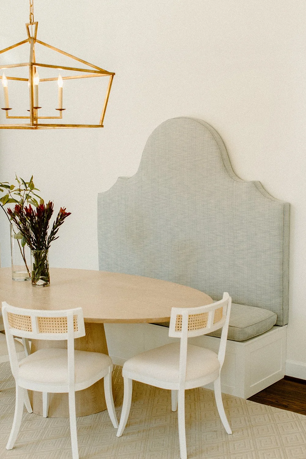 Dining nook with light gray cushioned bench, round wooden table, white chairs, gold geometric chandelier, and vase with flowers.