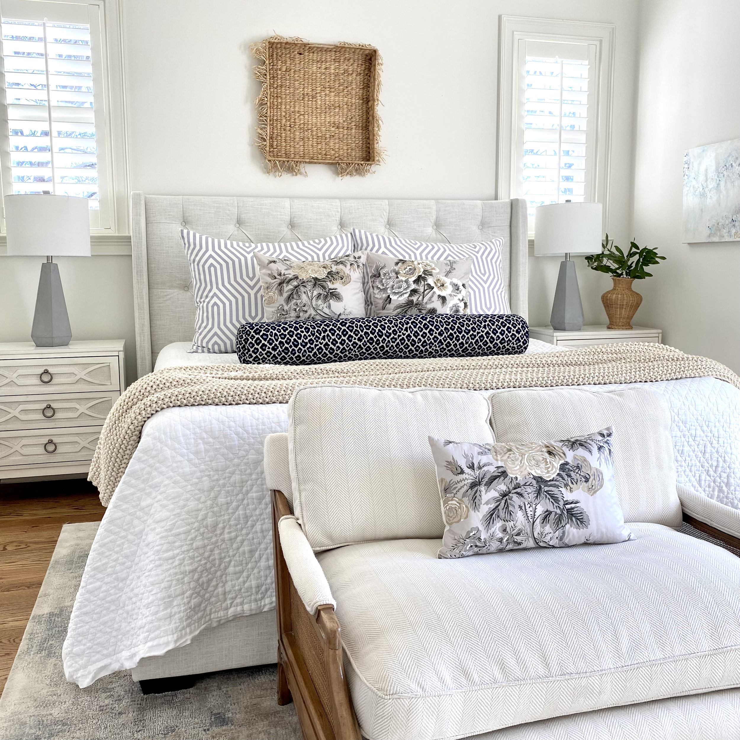 Modern bedroom with a white bed, decorative pillows, a beige knit blanket, two bedside tables with lamps, and a white armchair with a floral pillow. An empty wicker frame is mounted on the wall above the bed, and there are plants on both sides of the