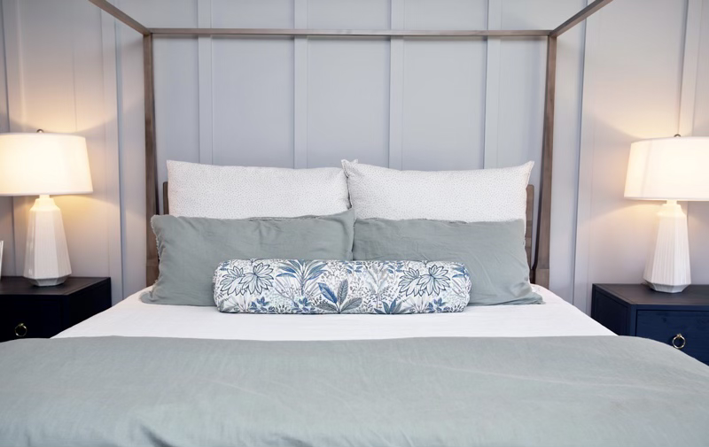 Stylish bedroom with a canopy bed, green bedding, patterned bolster pillow, white and gray pillows, and two nightstands with white lamps.