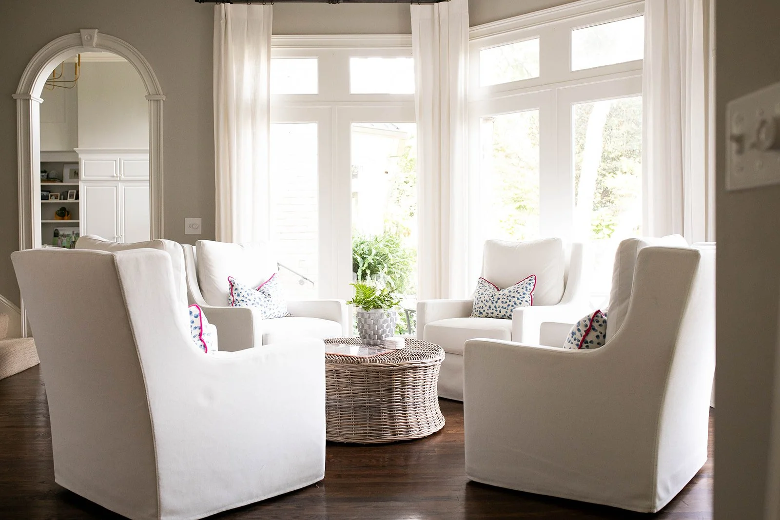 A bright living room with four white armchairs arranged in a circle around a wicker coffee table. The room features large windows with sheer curtains, wooden flooring, and decorative pillows with blue patterns on the chairs. A potted plant sits on th