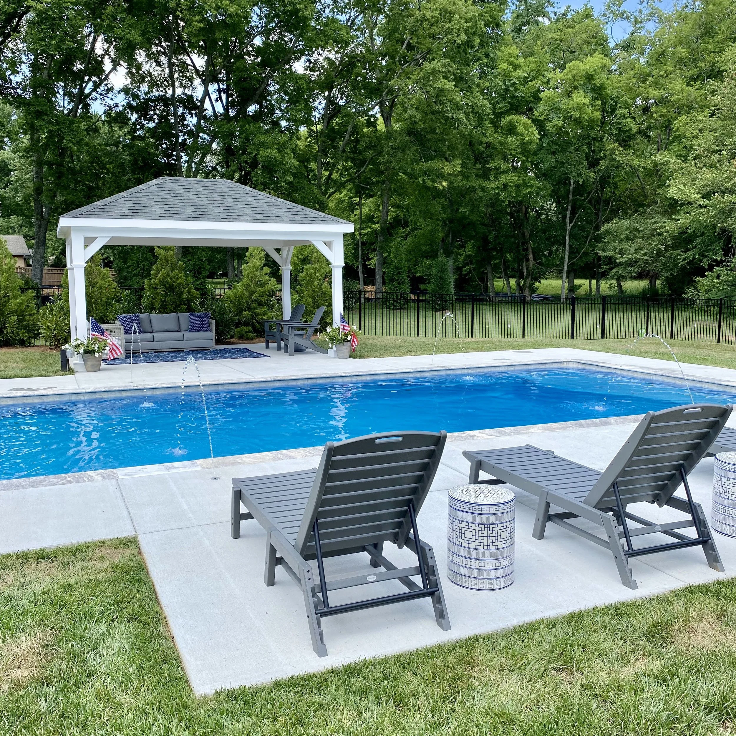 Backyard scene with a swimming pool, two lounge chairs, small tables, and a white gazebo in a garden setting.