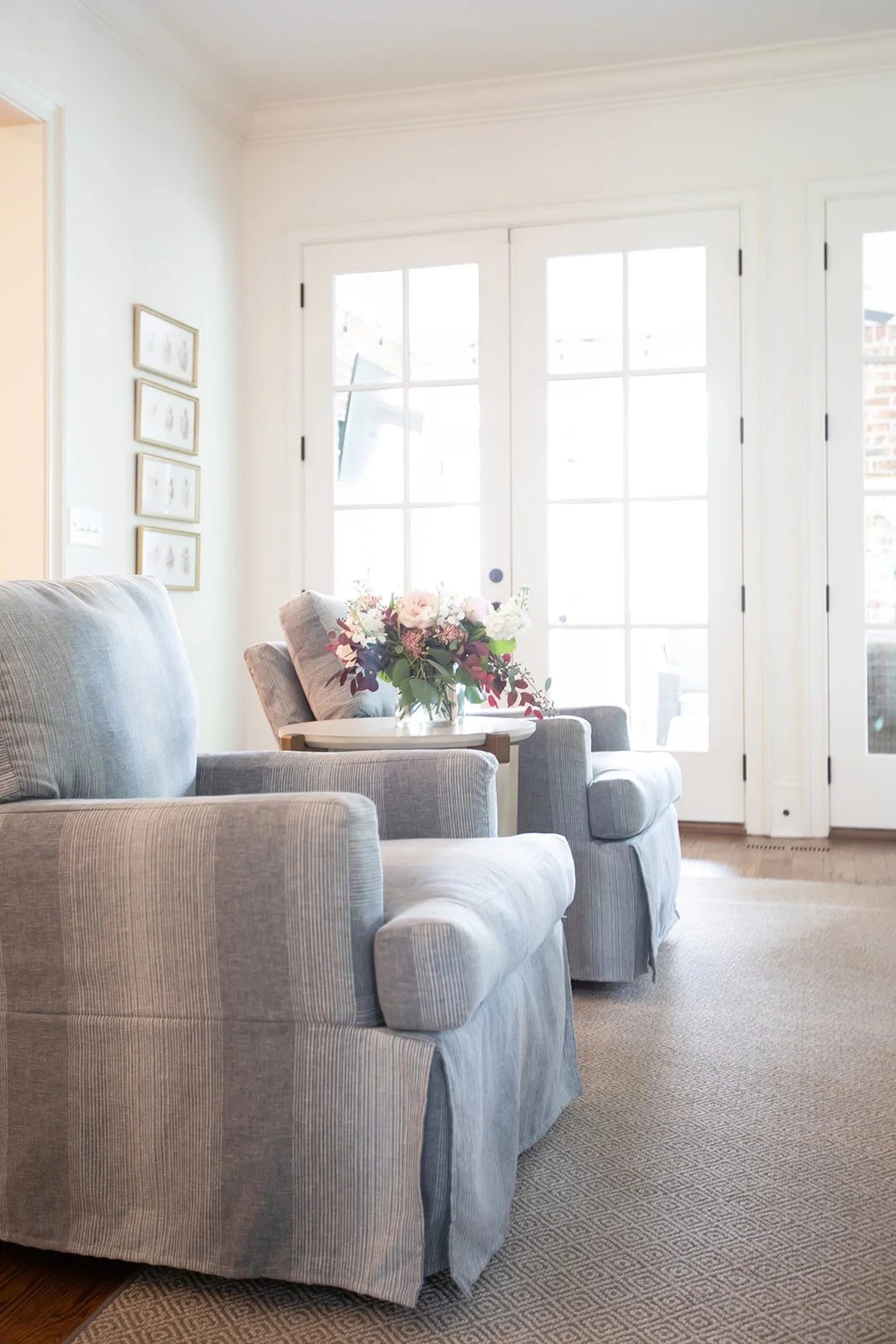 Bright living room with two gray armchairs, a floral arrangement on a side table, framed artwork on the wall, and large glass doors.