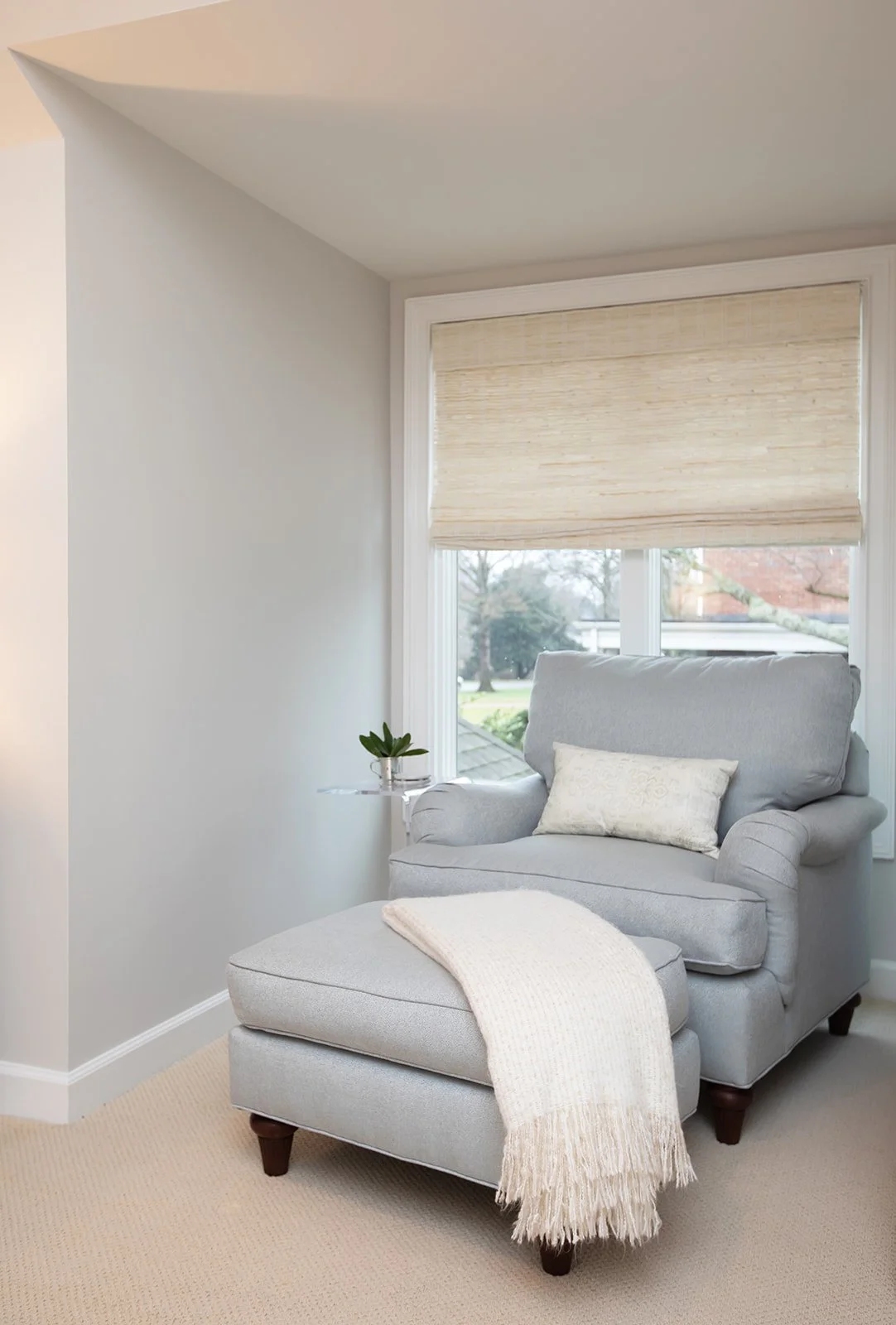 Cozy reading nook with gray armchair and matching ottoman, cream throw blanket, decorative pillow, small plant on side table, large window with blinds.