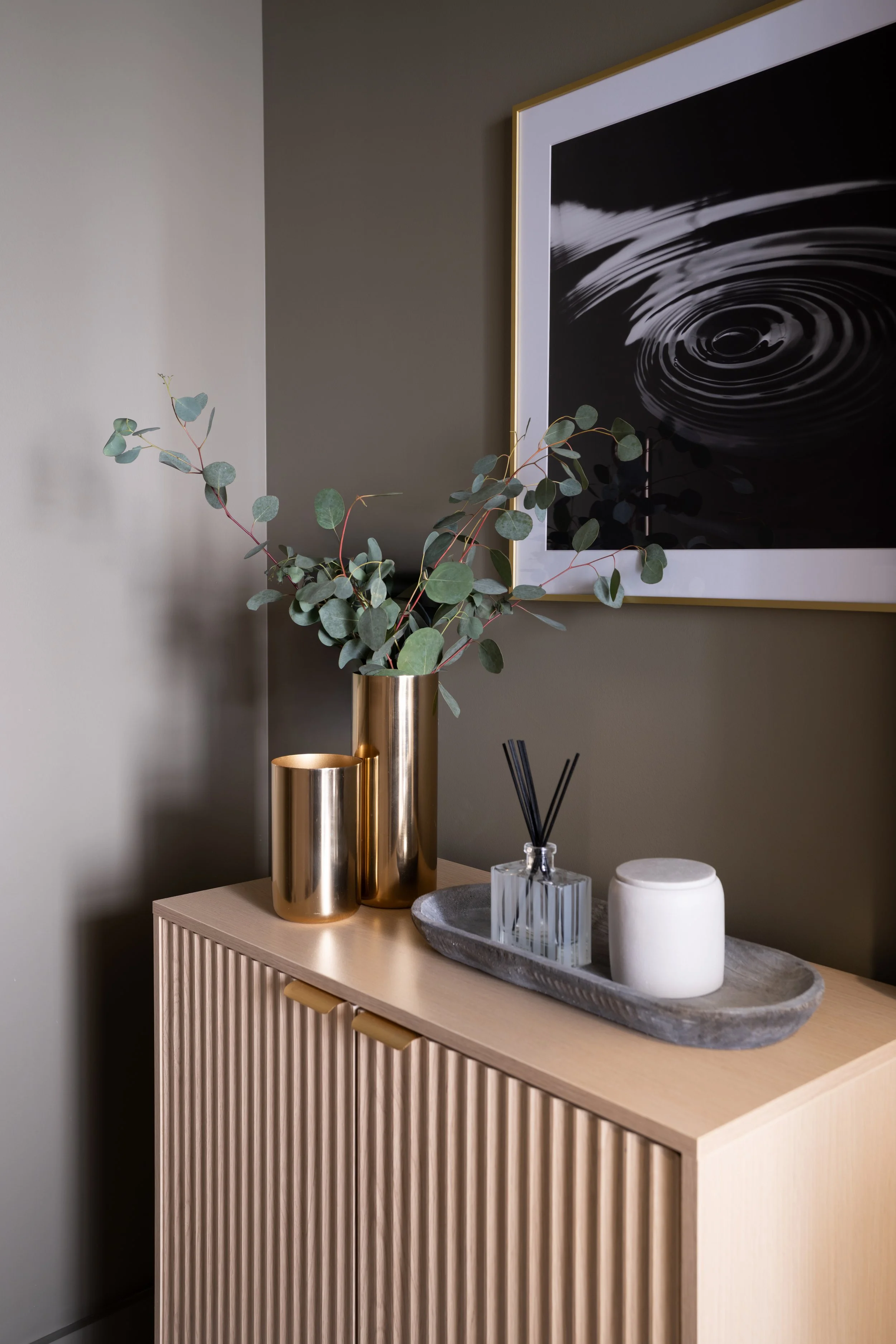 Decorative display on a light wood cabinet with green eucalyptus in gold vases, a tray with a diffuser, candle, and decorative container, against a dark green wall with black and white artwork.