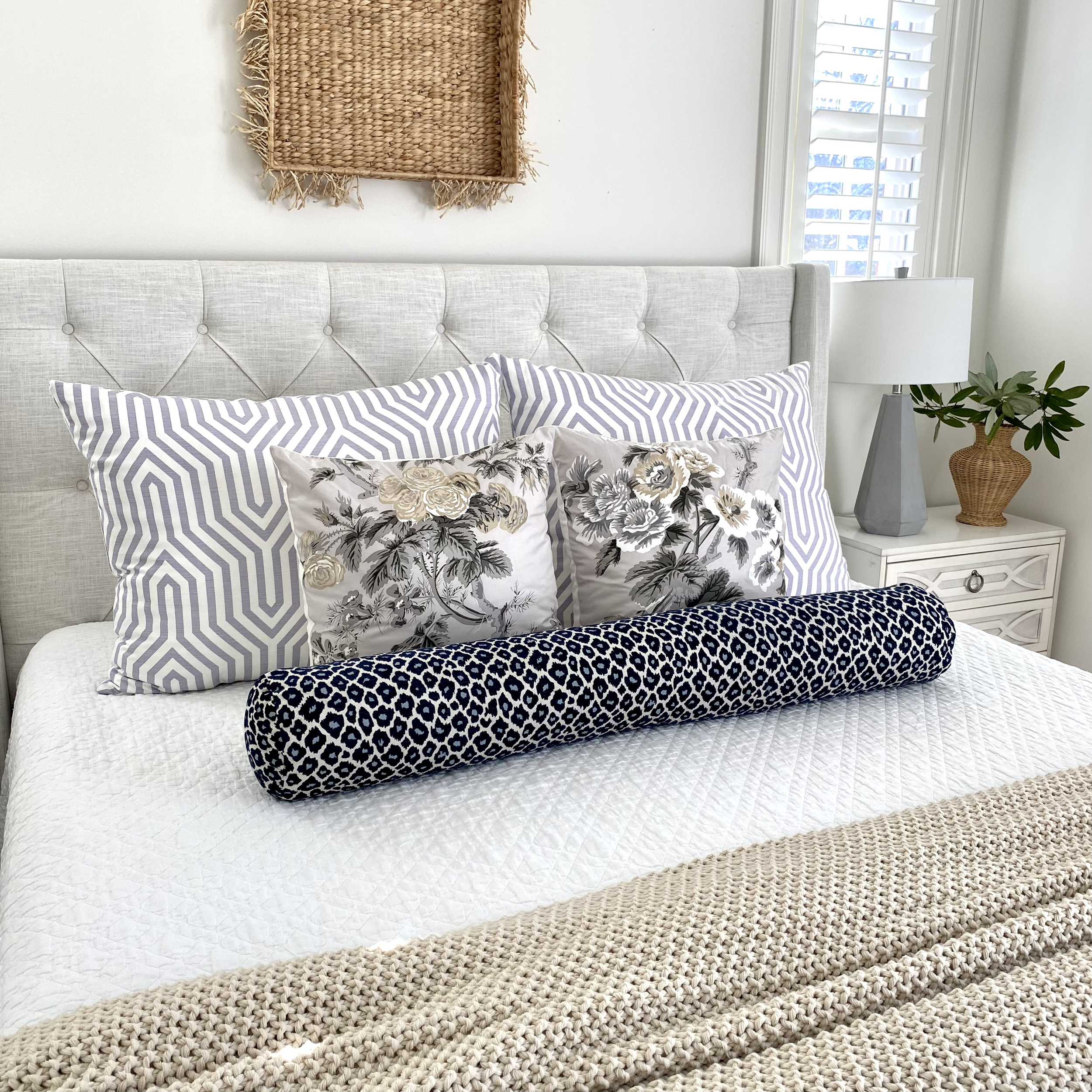A neatly made bed with a tufted headboard, featuring patterned pillows in blue, white, and floral designs. A navy bolster pillow is at the front. There is a beige knitted throw at the foot of the bed. A wicker basket serves as wall decor above the be