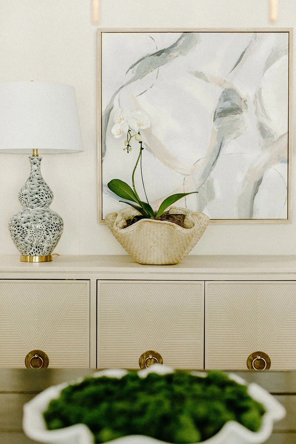 Interior scene with a decorative lamp, woven basket with an orchid, abstract painting, and a credenza with geometric patterns.