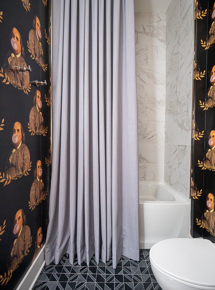 Stylish bathroom with black wallpaper featuring paintings of historical figures blowing bubble gum, a gray shower curtain, geometric black tile flooring, and a small bathtub.