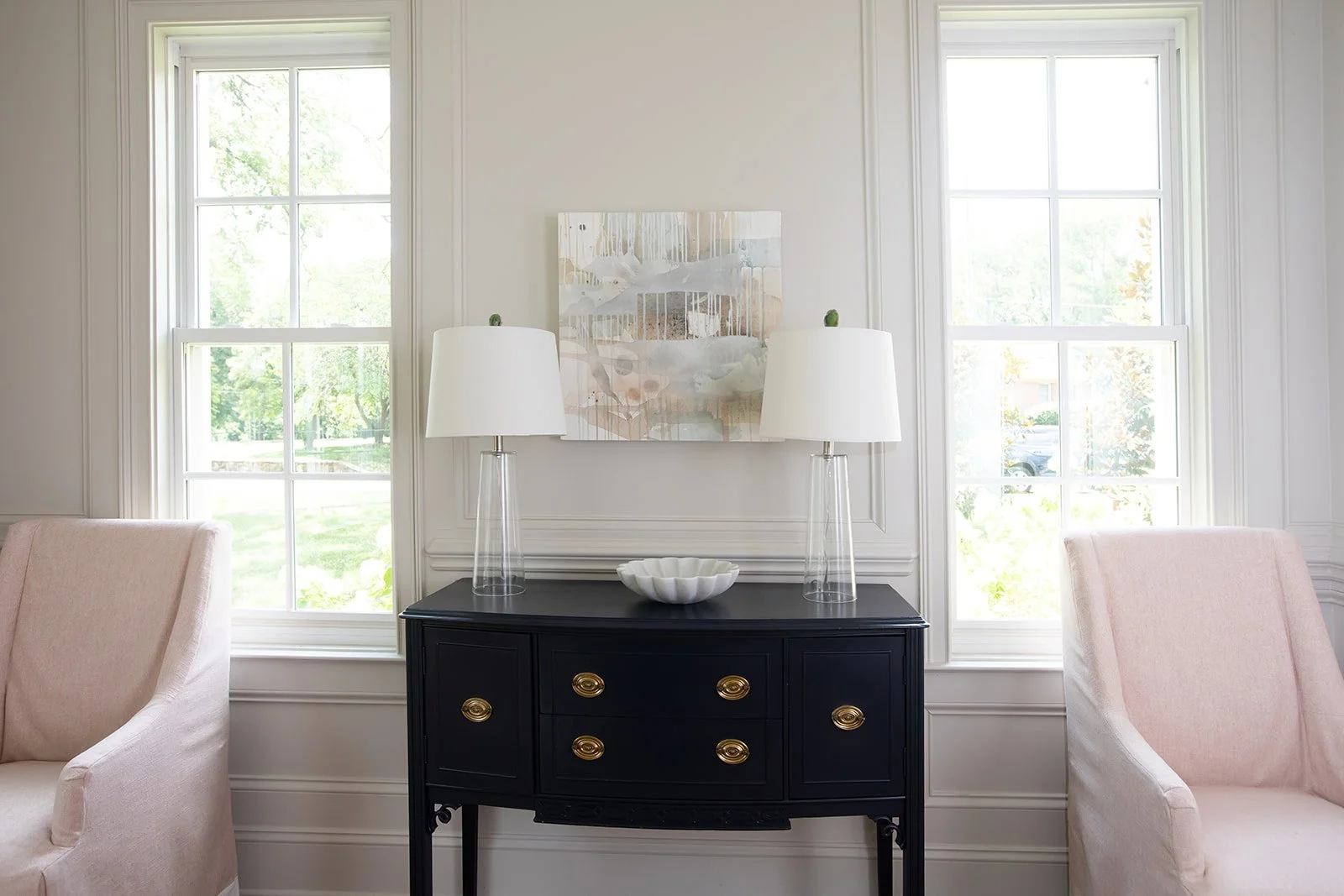 Interior living room with two armchairs, black sideboard, two table lamps, abstract wall art, and windows.