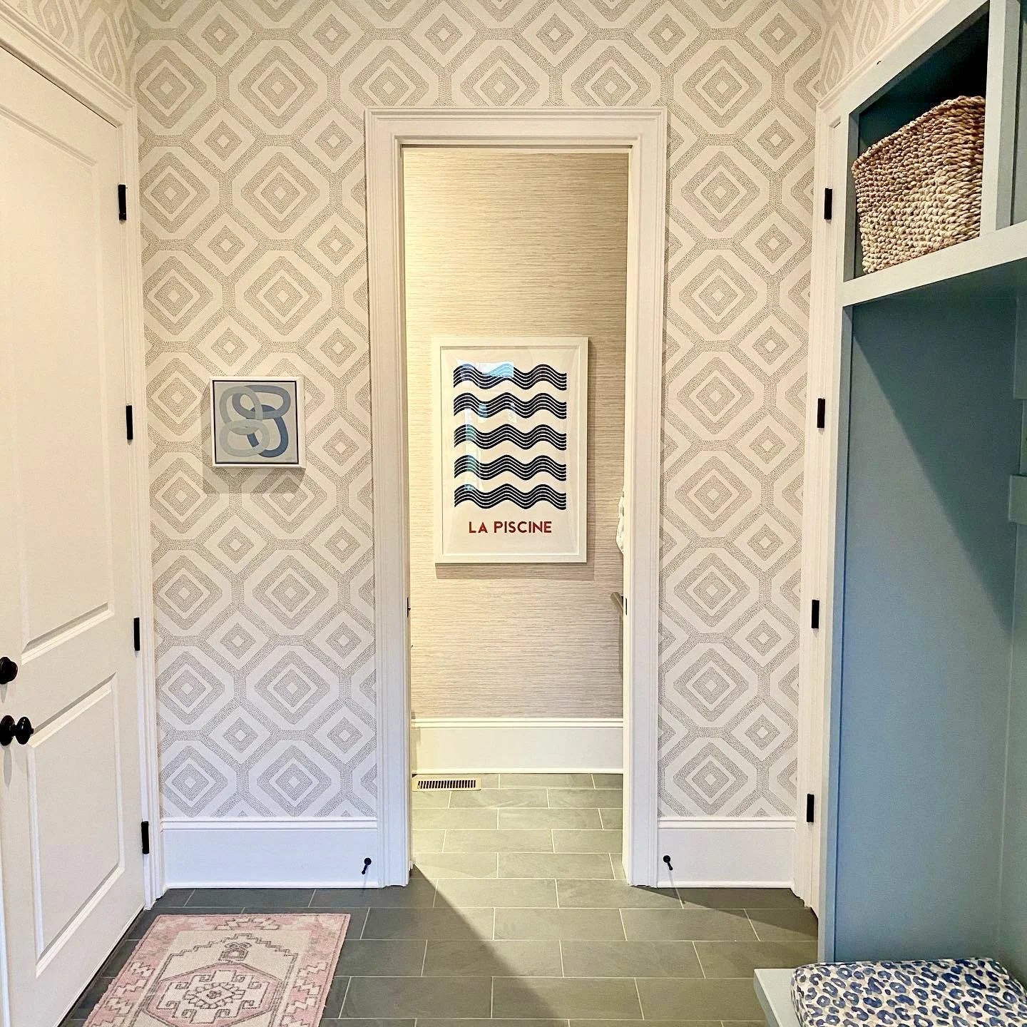 Interior hallway with geometric wallpaper, a rug, and artwork on the wall labeled "La Piscine." There's a door on the side and a shelf with a basket.