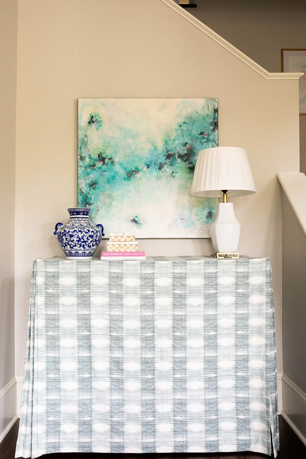 Table with blue and white ceramic vase, books, white lamp, abstract painting, and checkered tablecloth in a hallway.