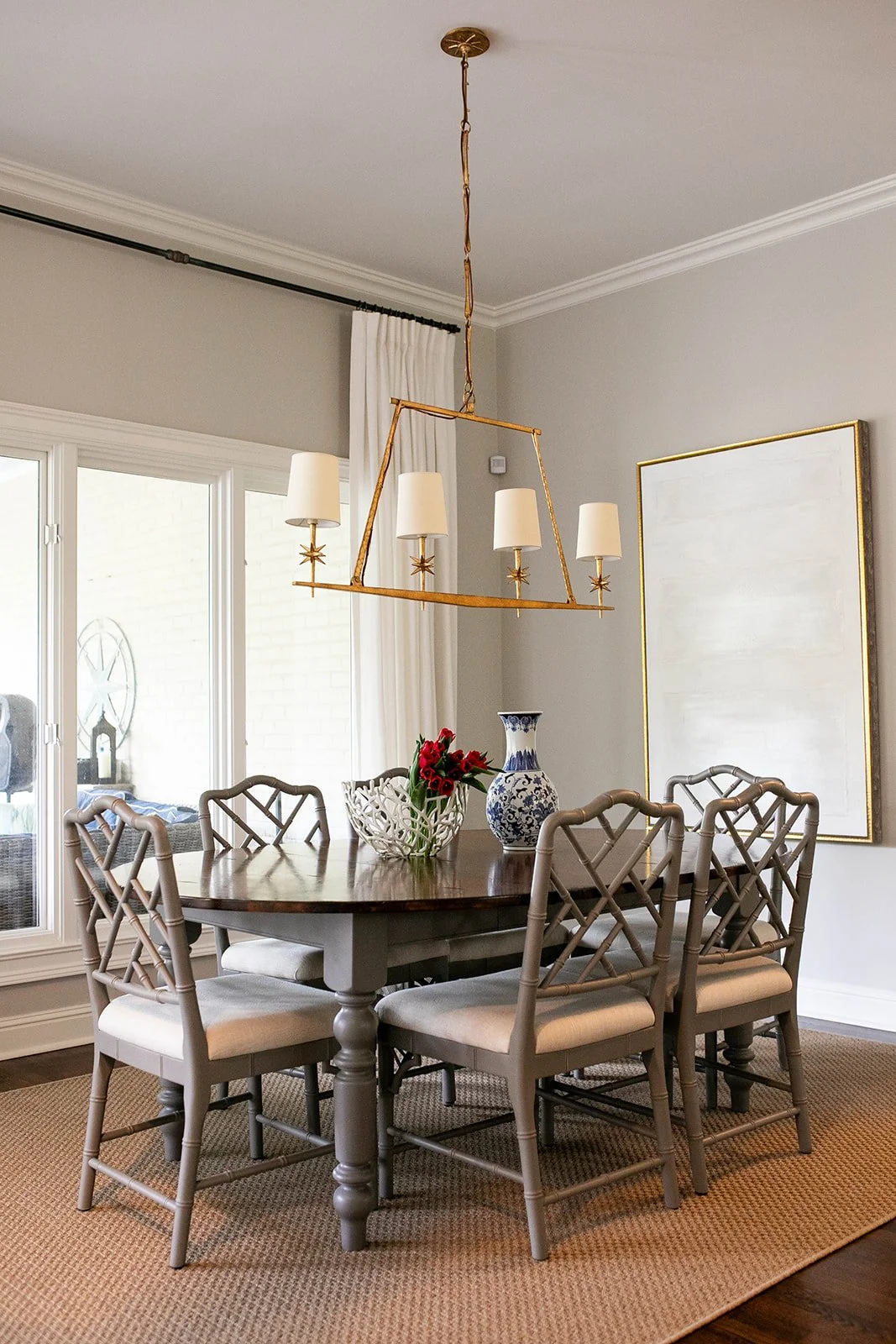 Dining room with wooden table, six chairs, chandelier, vase with red flowers, blue and white decorative vase, neutral artwork, large windows with curtains, and beige carpet.