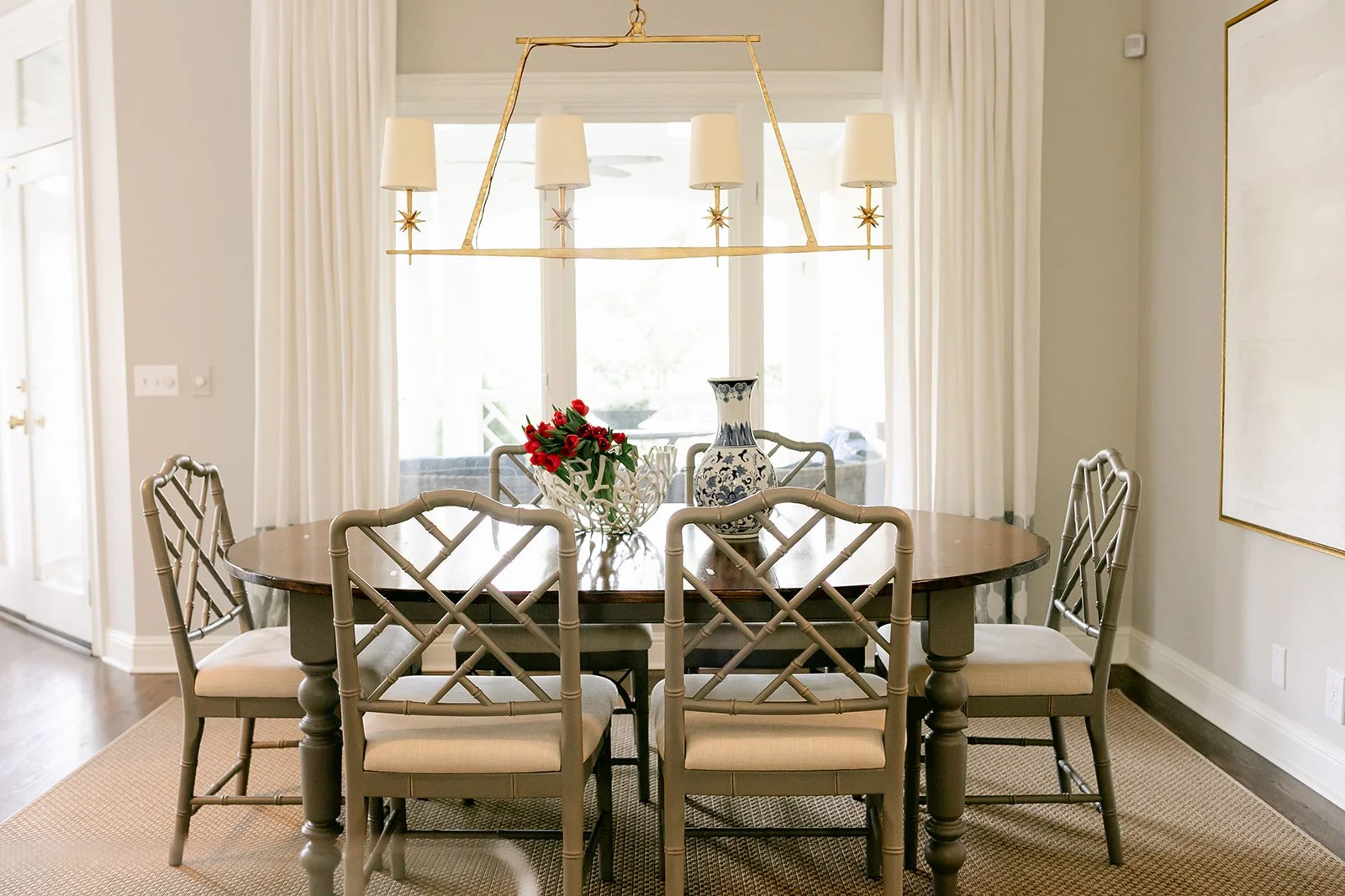 Elegant dining room with round wooden table, six gray chairs, gold chandelier, and decorations including a blue patterned vase and red flowers.