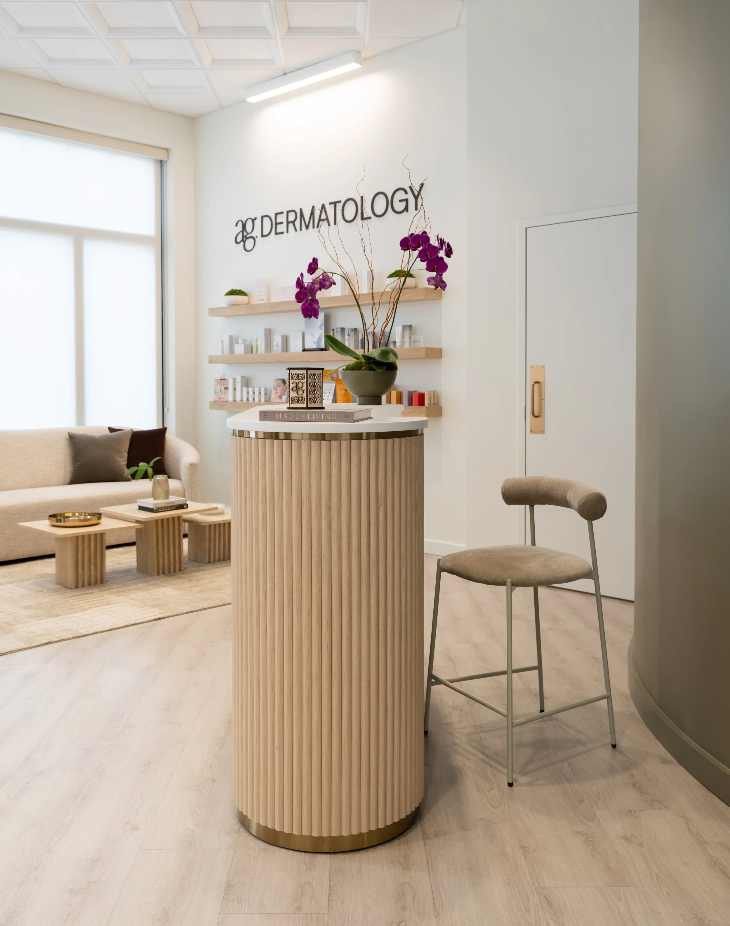 Interior view of a dermatology clinic waiting area with a beige plush sofa, wooden coffee tables, decorative books, and shelves with skincare products. A round, fluted wooden reception desk with a potted purple orchid on top is in the foreground. The