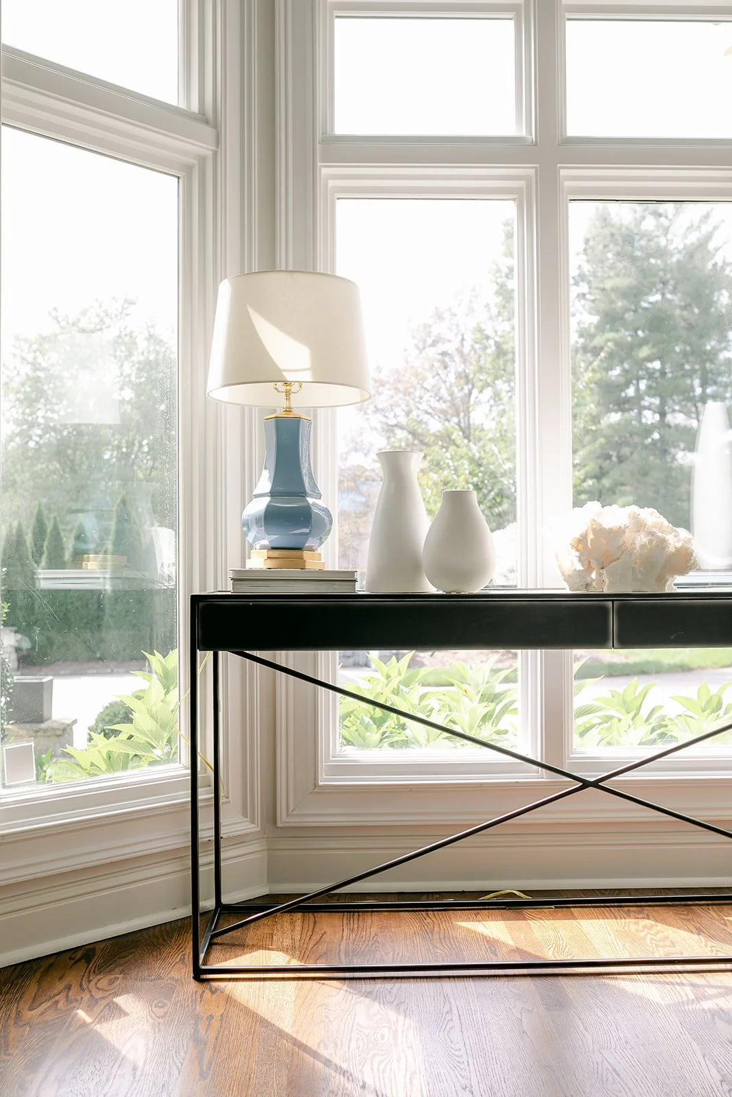 A modern room with large windows featuring a white lamp with a blue base on a black table, two white vases, and a piece of coral sculpture. Sunlight filters in, illuminating the hardwood floor and greenery outside.