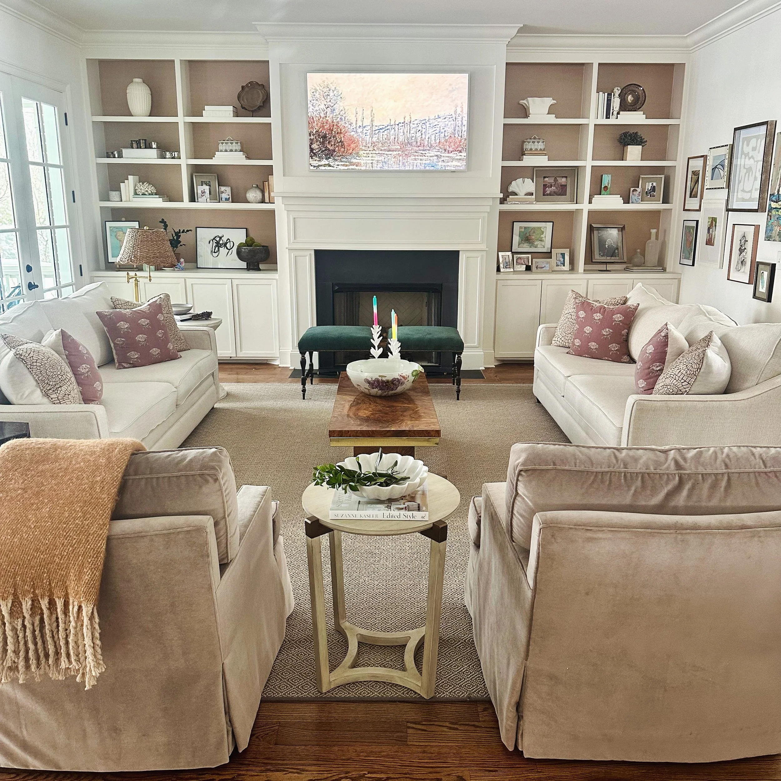 Living room with white sofas, pink pillows, fireplace, and wall shelves. Coffee table with decorative items and green cushioned bench. Artwork above the fireplace, framed pictures on walls. Soft lighting and neutral tones.