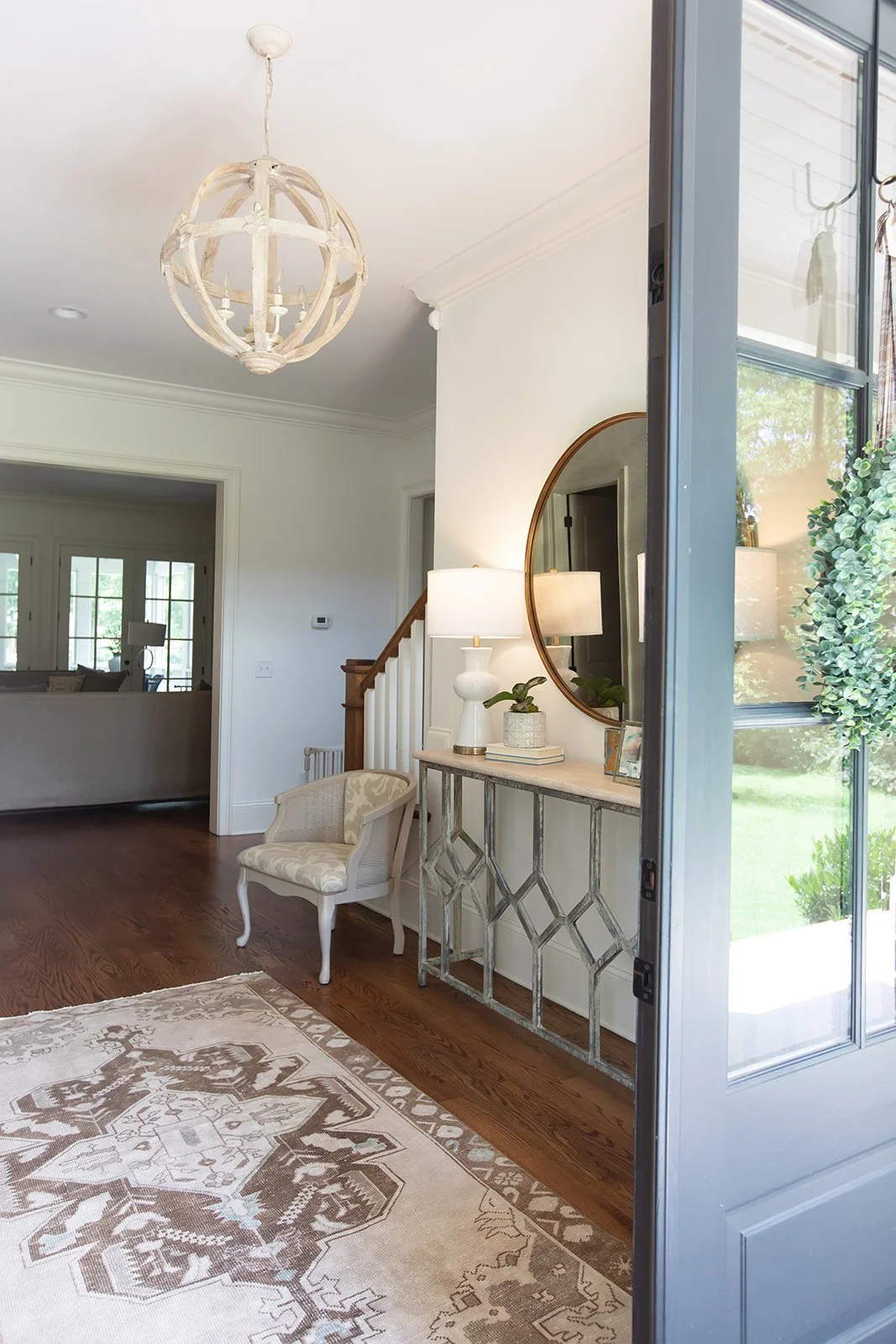 Bright, elegant foyer with wooden flooring, decorative patterned rug, entry table with plants and lamp, round mirror above table, armchair, spherical chandelier, and open door leading outside.