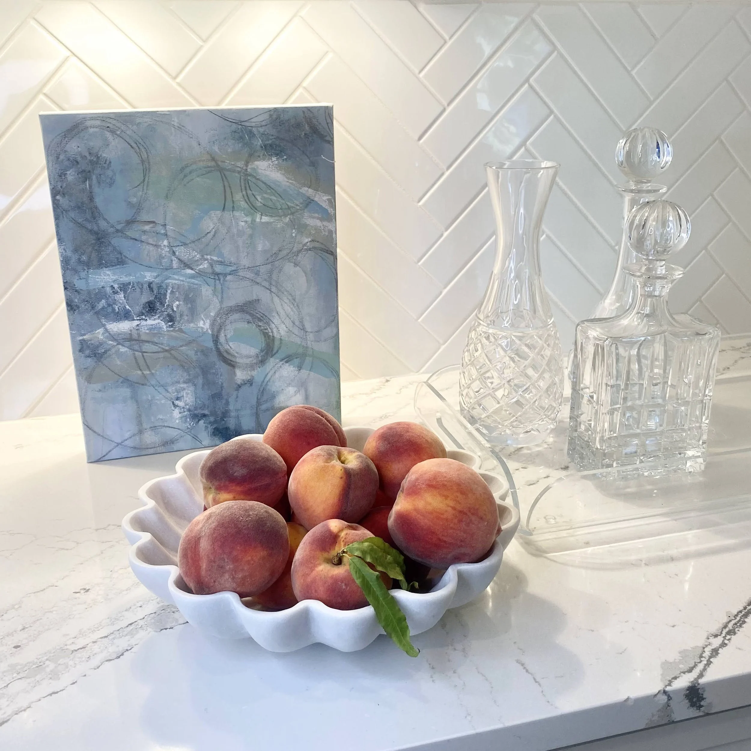 A bowl of peaches on a marble countertop next to a blue abstract painting and two crystal decanters, against a white tiled backsplash.