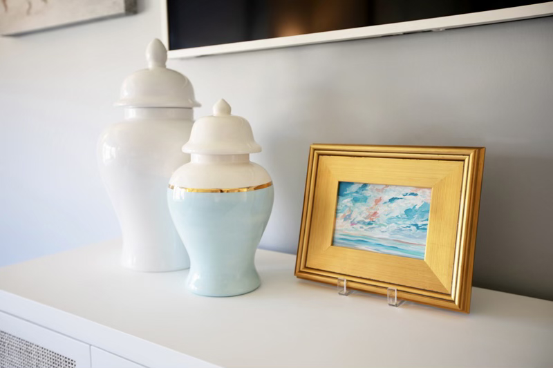 A white shelf displaying two decorative jars and a framed painting. The jars are lidded, one white and the other with a light blue accent and gold trim. The painting has a gold frame and shows an abstract landscape.