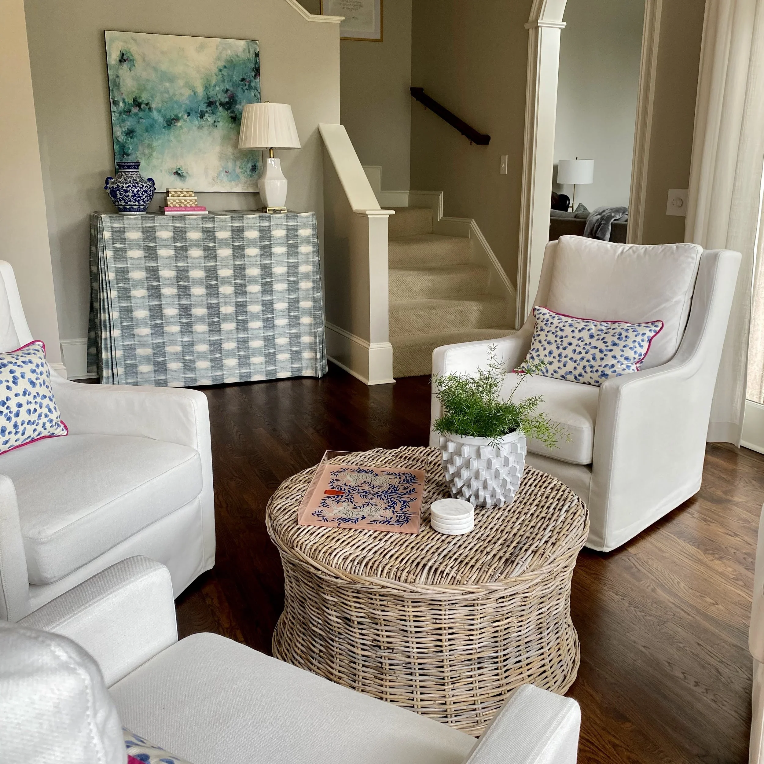 Stylish living room with white armchairs, patterned pillows, a wicker coffee table with a plant, and a decorative console with art and a lamp.