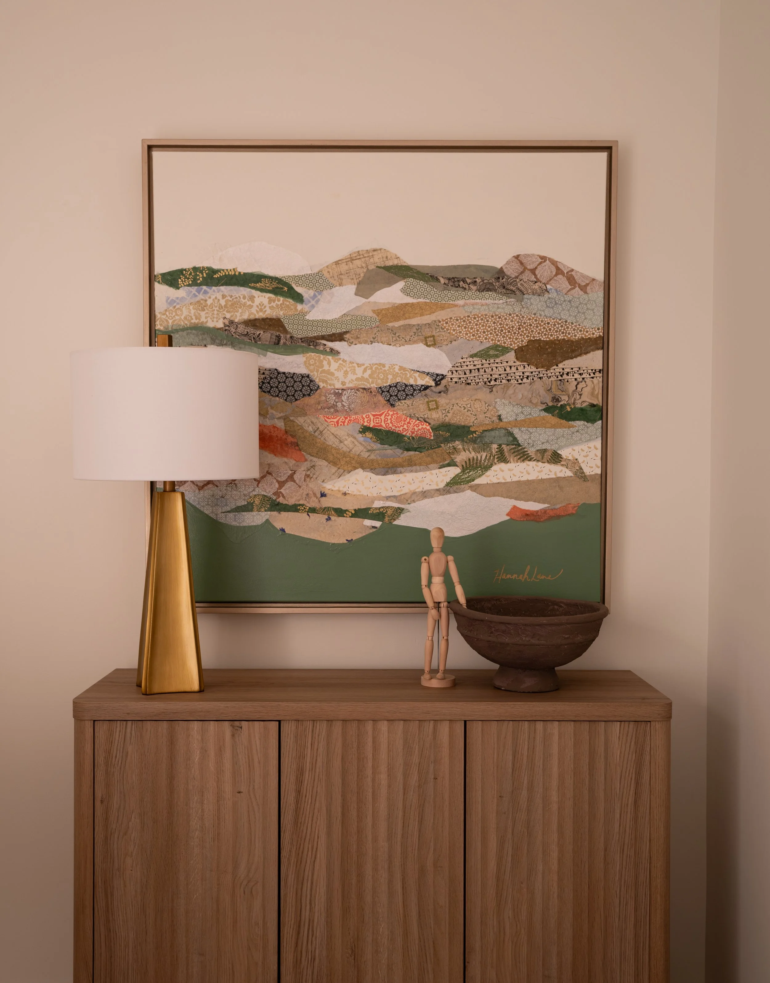 A wooden sideboard with a white lampshade with a gold base, a wooden artist mannequin, and a large brown bowl. A framed abstract landscape painting with layered patterns and colors hangs on the wall behind.