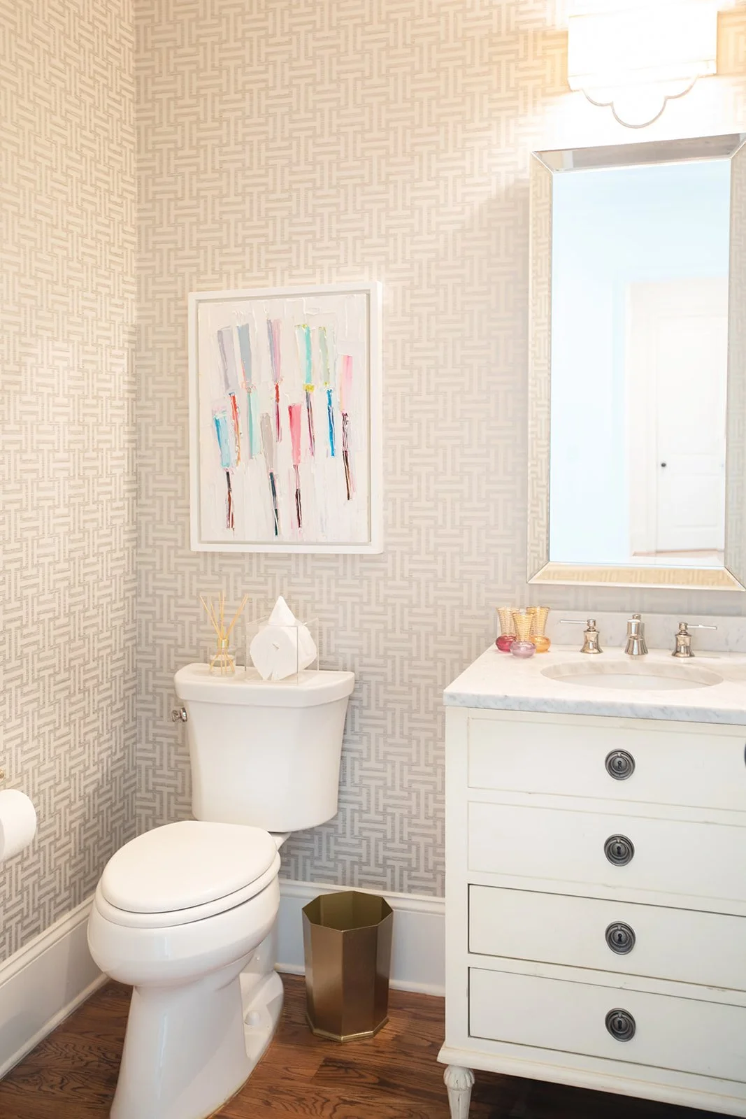 A bathroom with patterned wallpaper, a toilet, a wastebasket, a countertop with a sink and decorative items, and a large mirror. Artwork with colorful brushstrokes is hung on the wall.
