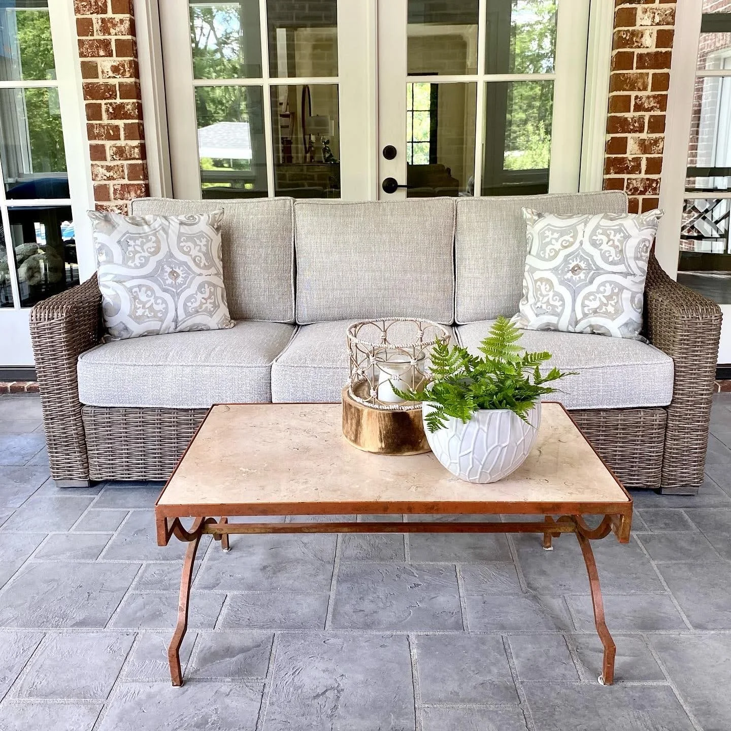 Patio with a wicker sofa featuring gray cushions and decorative pillows, a stone-top coffee table with a potted plant and decorative items, in front of glass doors and brick walls.
