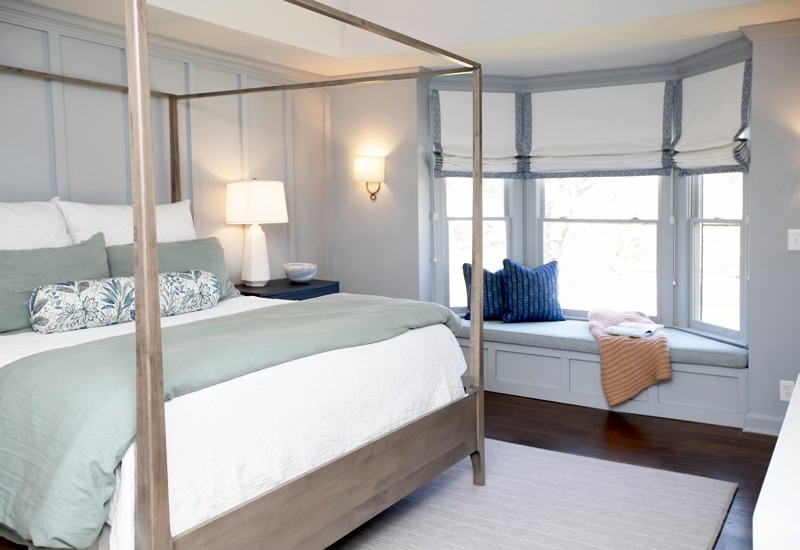 Modern bedroom with a canopy bed, light blue bedding, and decorative pillows. There's a bedside table with a lamp, a window seat with cushions and a throw blanket, and gray wall paneling. The room has a soft, serene ambiance.