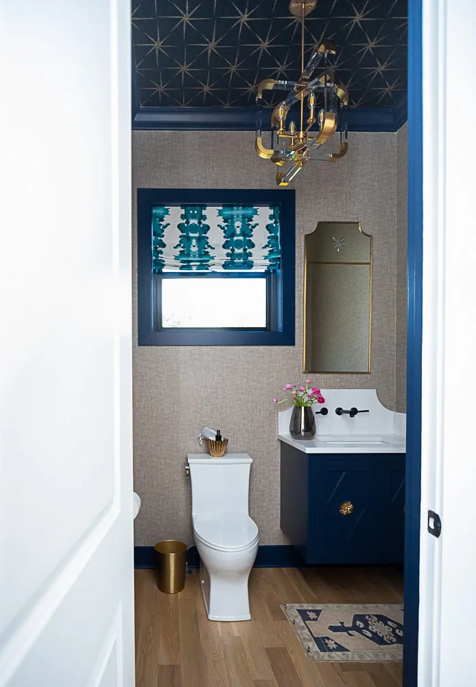 Stylish modern bathroom with blue accents, featuring a white toilet, blue vanity, decorative mirror, and floral arrangement. Gold chandelier, geometric ceiling, patterned window treatment, wooden floor, and small rug complete the design.