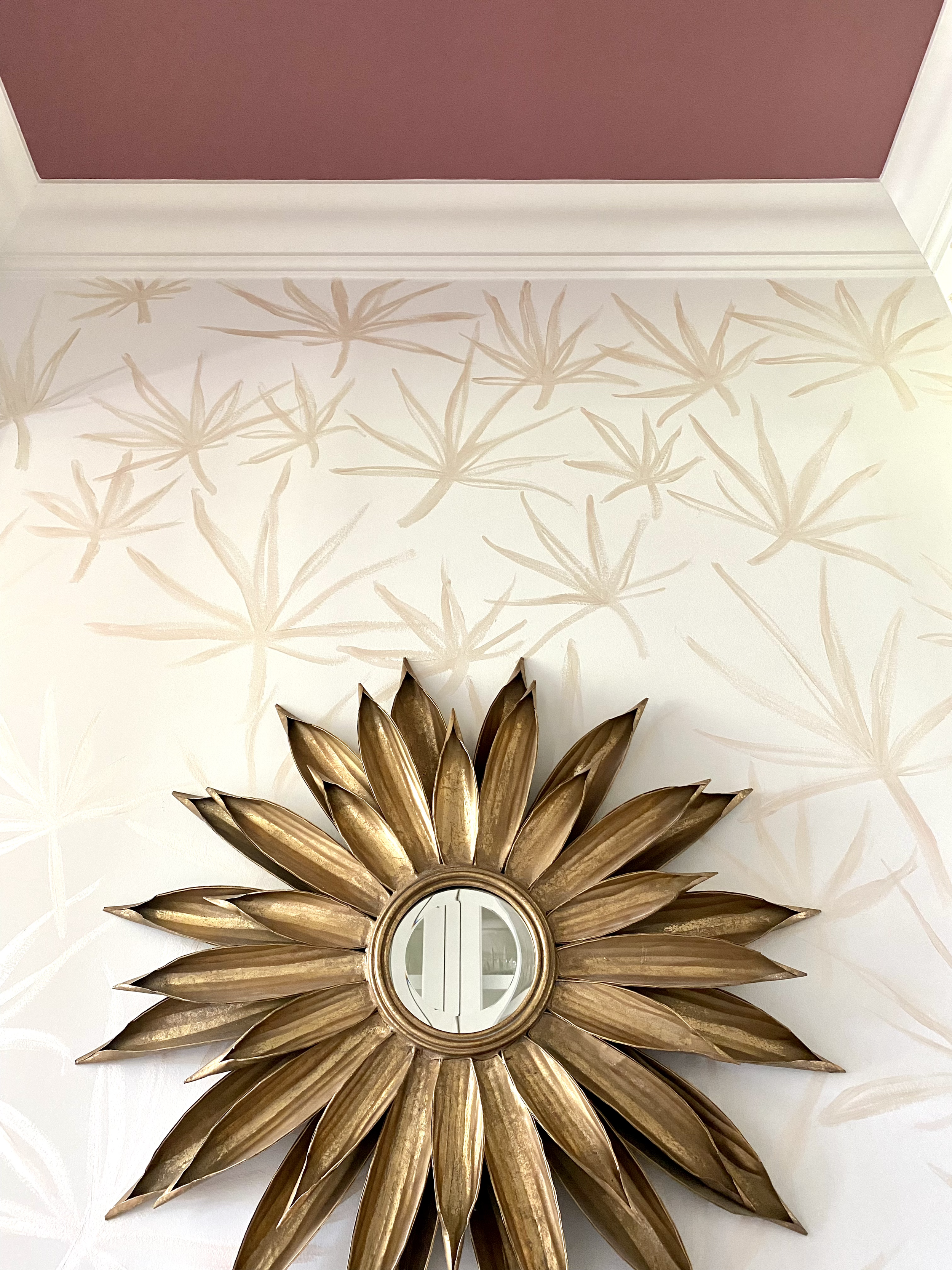 Decorative wall with gold starburst mirror and leaf pattern wallpaper beneath a maroon ceiling.