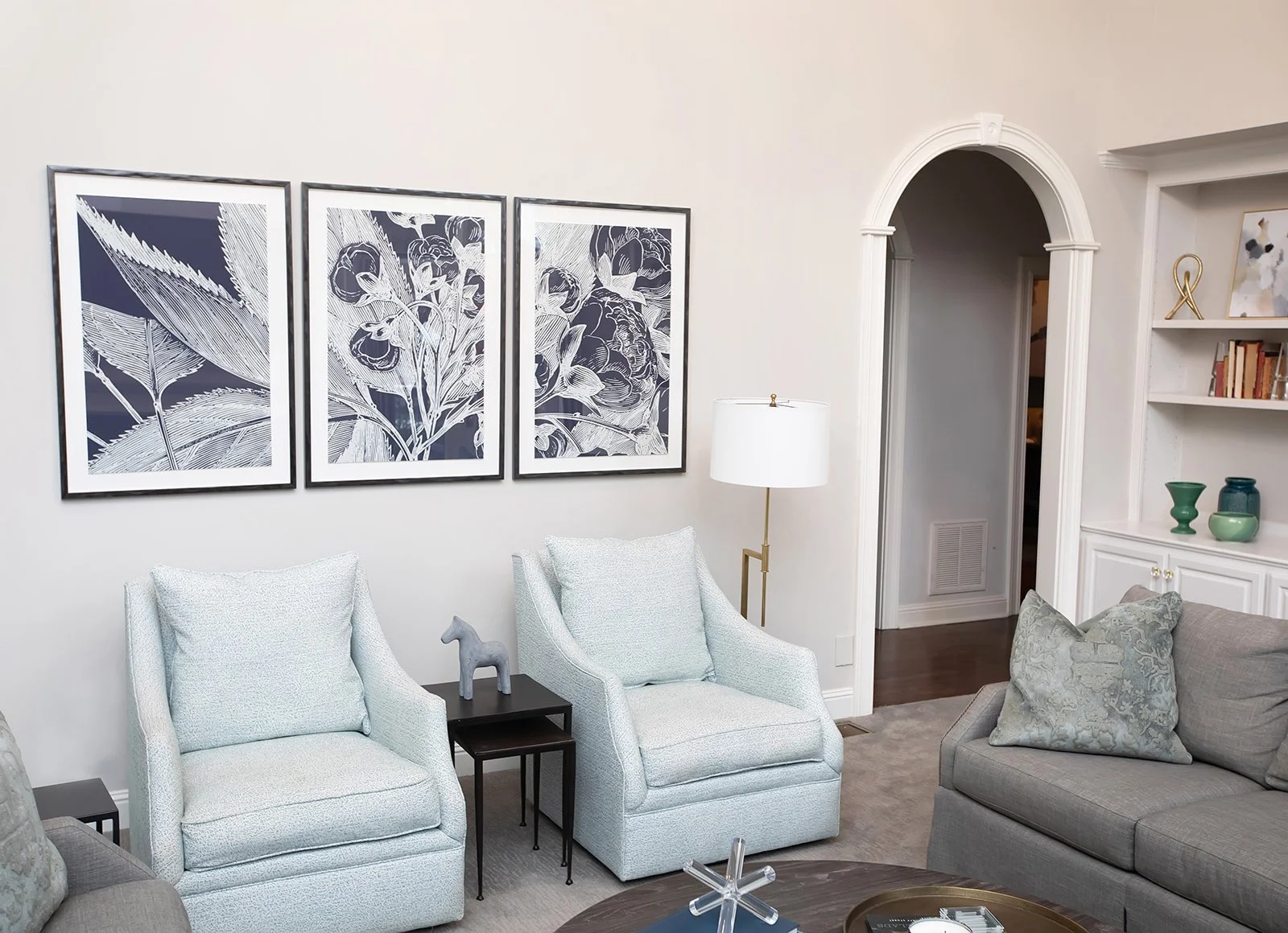 Living room with two light blue armchairs, three framed botanical prints on the wall, a floor lamp, a gray sofa with a patterned pillow, and bookshelves with decorative items.