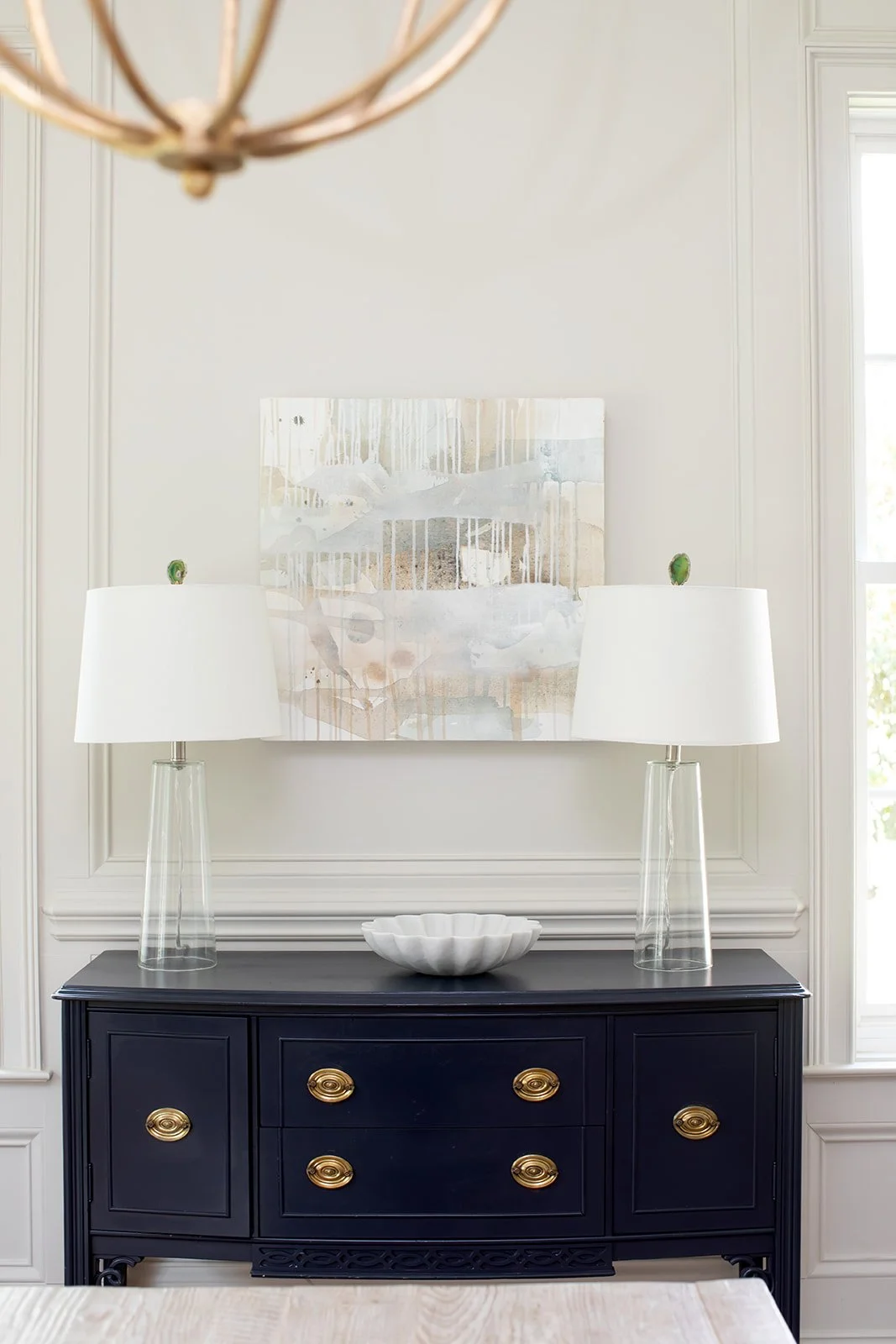 Elegant interior decor featuring a black sideboard with gold handles, topped with two glass-based lamps with white shades and a white decorative bowl. Abstract artwork hangs on the wall above, and a light fixture is partially visible.