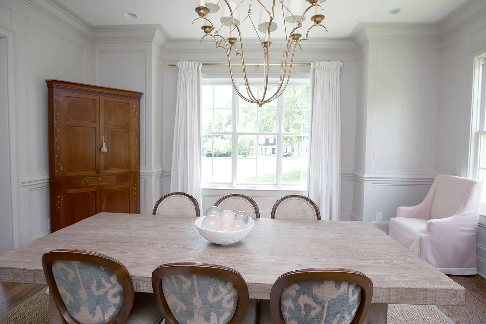 Dining room with a wooden table, decorative chairs, chandelier, large window with white curtains, a wooden cabinet, and an upholstered armchair.