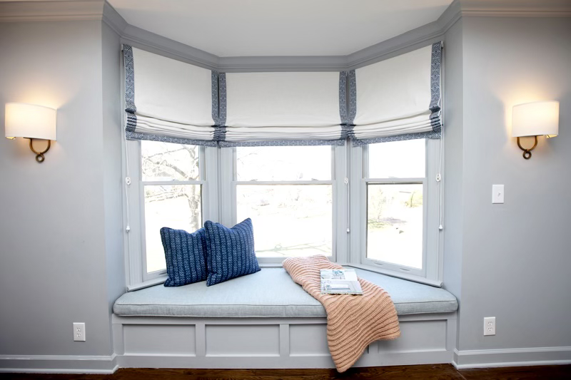 Cozy window seat with blue cushions, beige blanket, and reading material in a bright room with white roman shades and wall sconces.