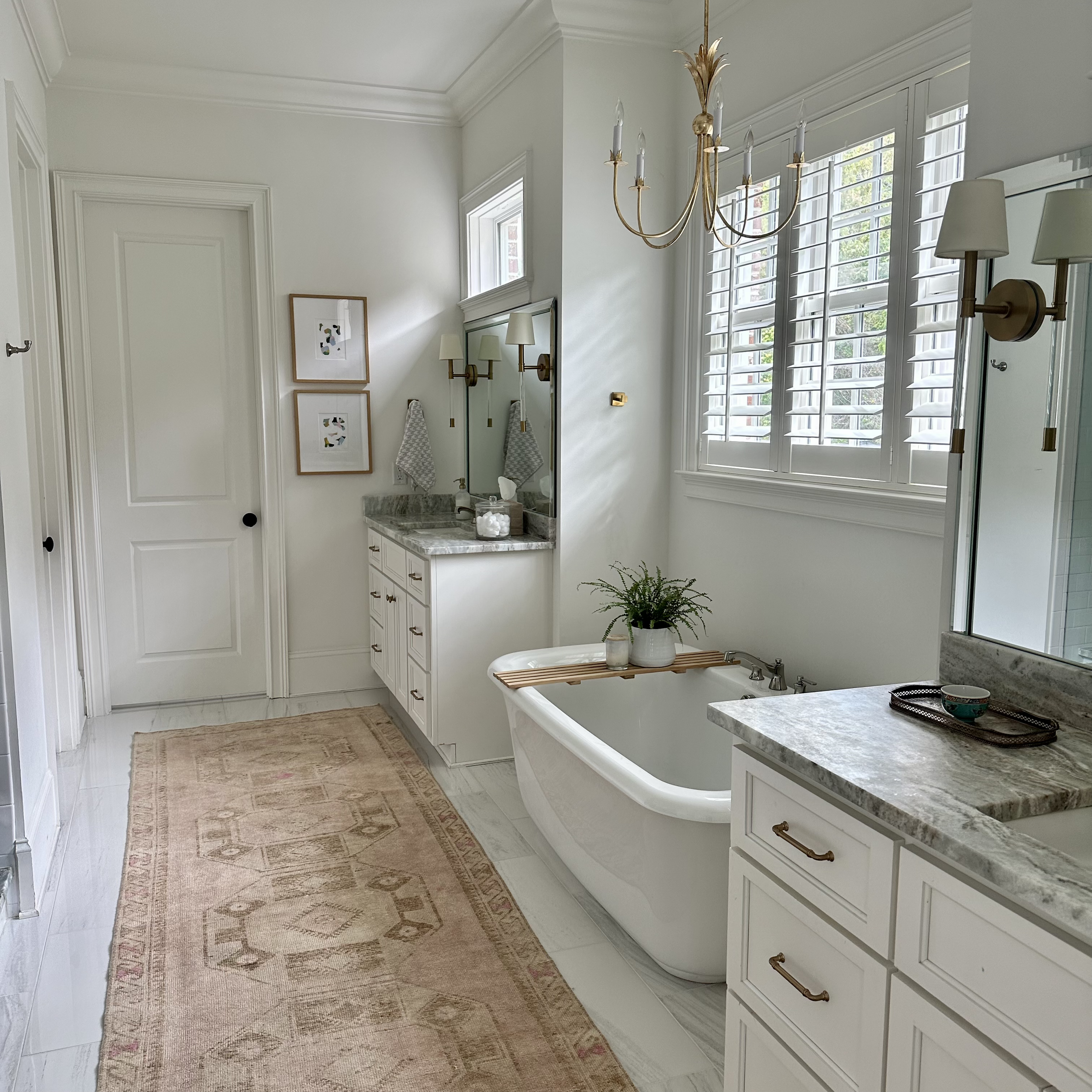 Elegant bathroom with white cabinets, gray marble countertops, a freestanding bathtub with a wooden tray and potted plant, a chandelier, wall-mounted lamps, framed art, a runner rug, and window shutters.