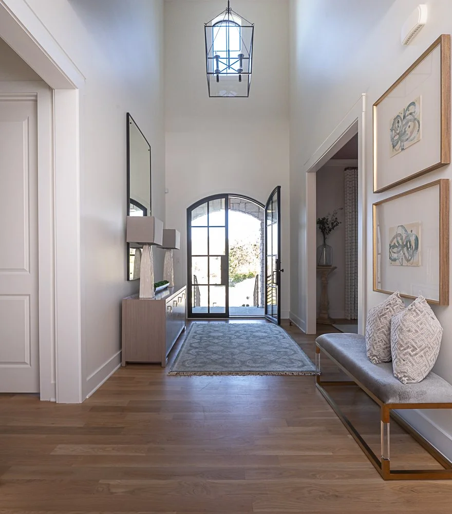 Modern entryway with wooden floor, large glass door, bench with cushions, wall art, console table with lamps, and a chandelier.