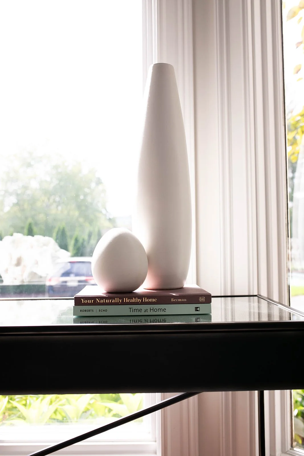 A tall white ceramic vase and a smaller round ceramic sculpture on a glass table, with books underneath them near a bright window.