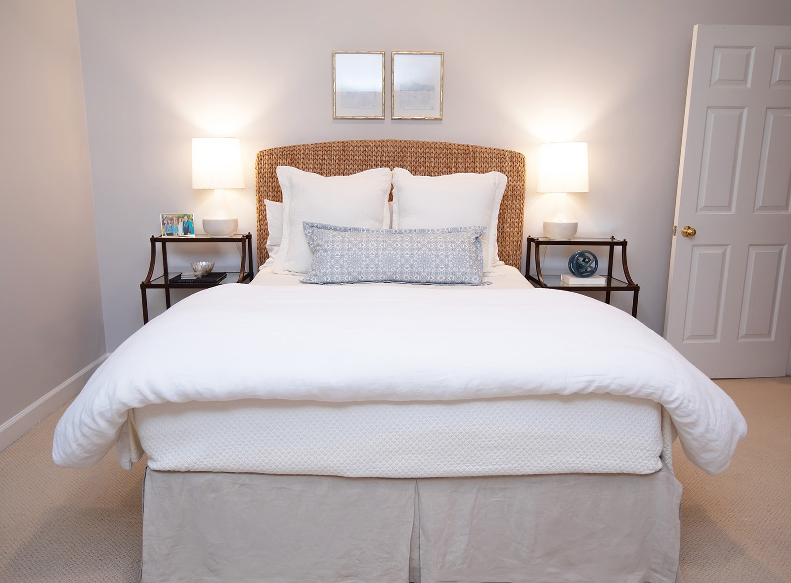 Contemporary bedroom with a neatly made bed, white bedding, wicker headboard, two bedside tables with lamps, and framed art above the bed.