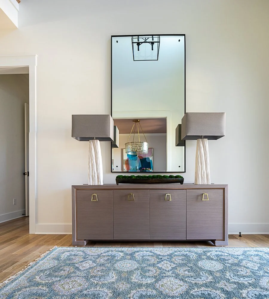 Modern living room interior with a large mirror above a wooden sideboard, two large table lamps with rectangular shades, and a decorative center piece. The room has a light-colored wall and hardwood flooring with a patterned area rug.